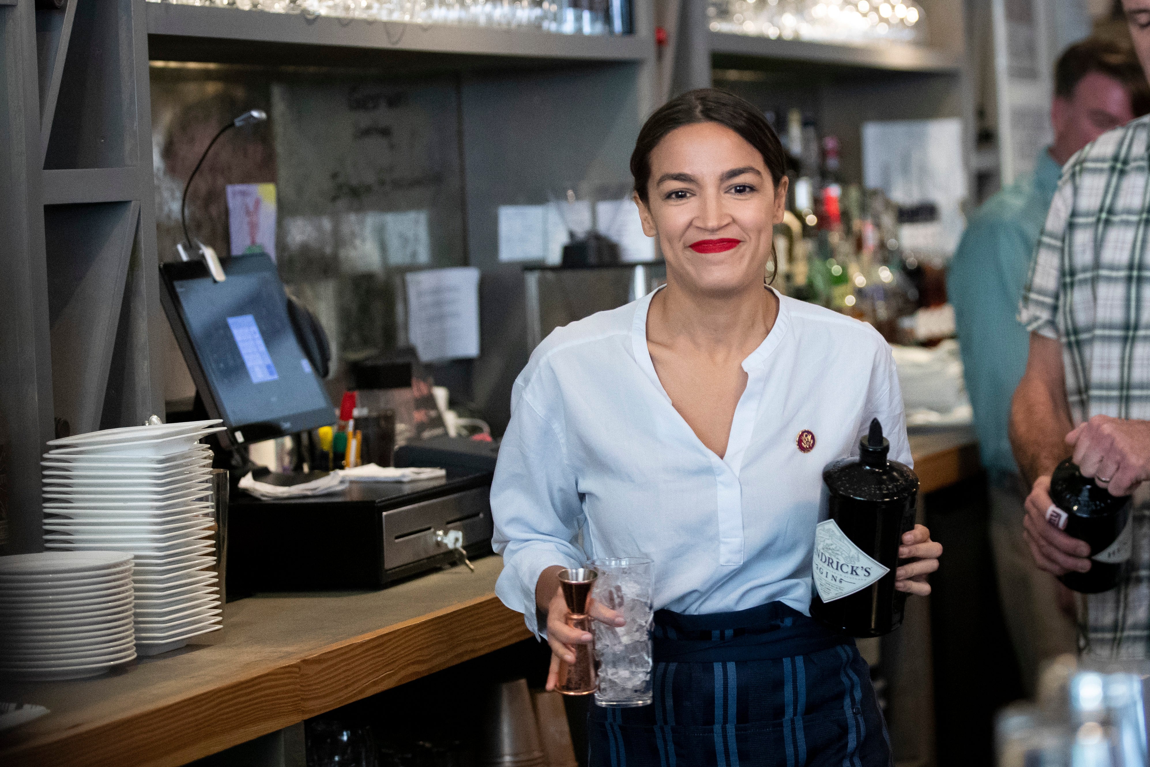 Person down a bar, smiling, holding a portion and a bottle, wearing a achromatic blouse and striped apron