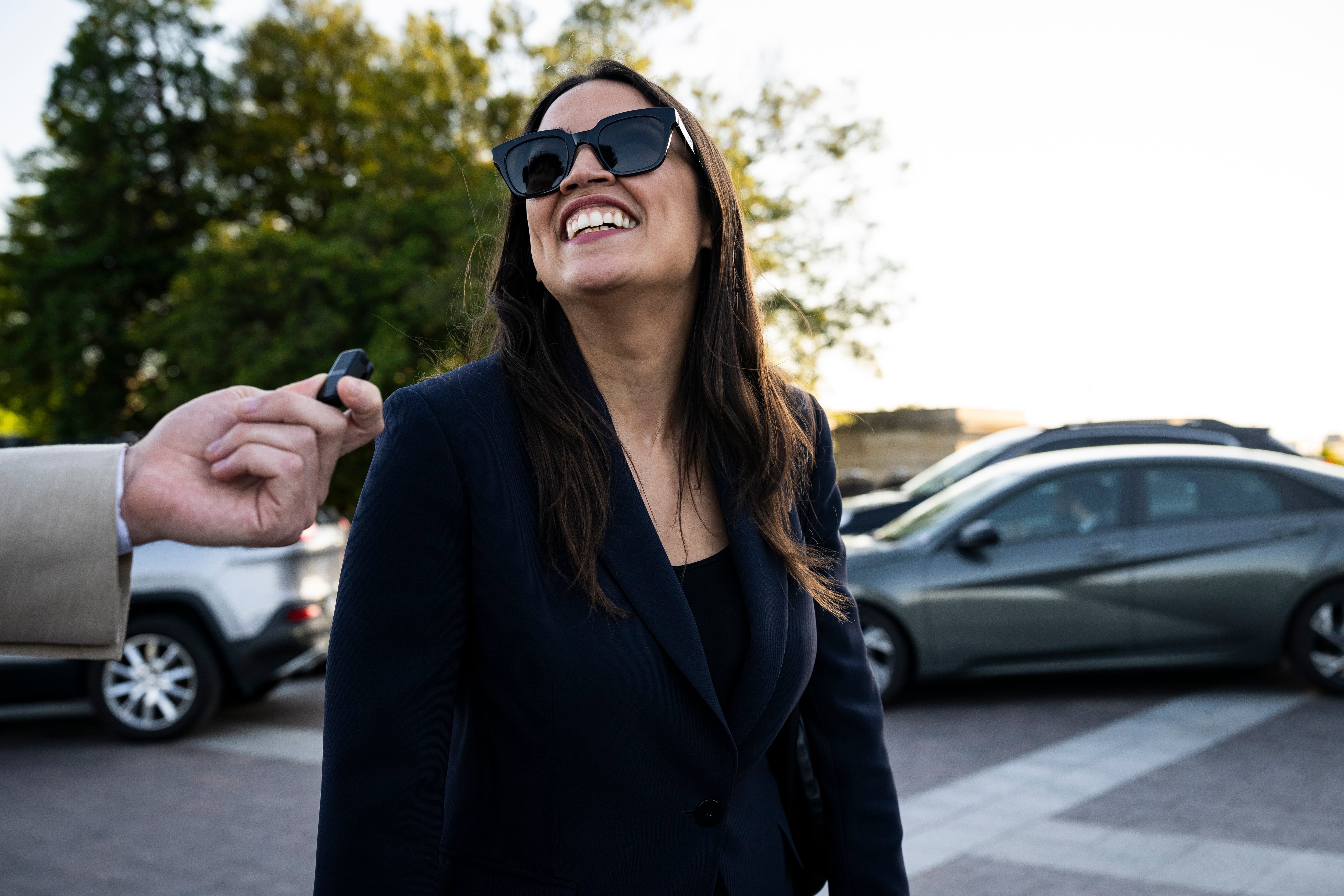 A idiosyncratic smiling outdoors, wearing acheronian sunglasses and a stylish acheronian blazer, portion idiosyncratic holds a microphone nearby