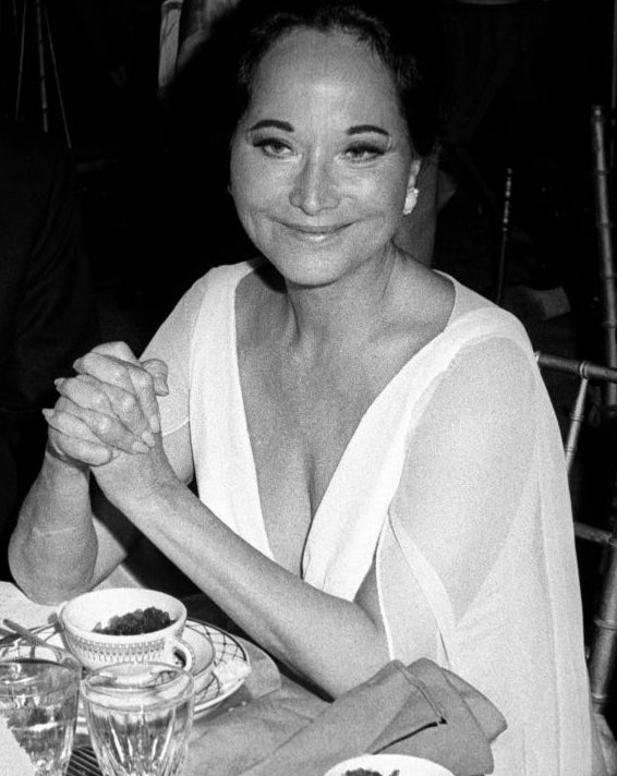 A idiosyncratic   successful  ceremonial  attire is seated astatine  a table, smiling with clasped hands, surrounded by dishes and glasses astatine  what appears to beryllium  an event