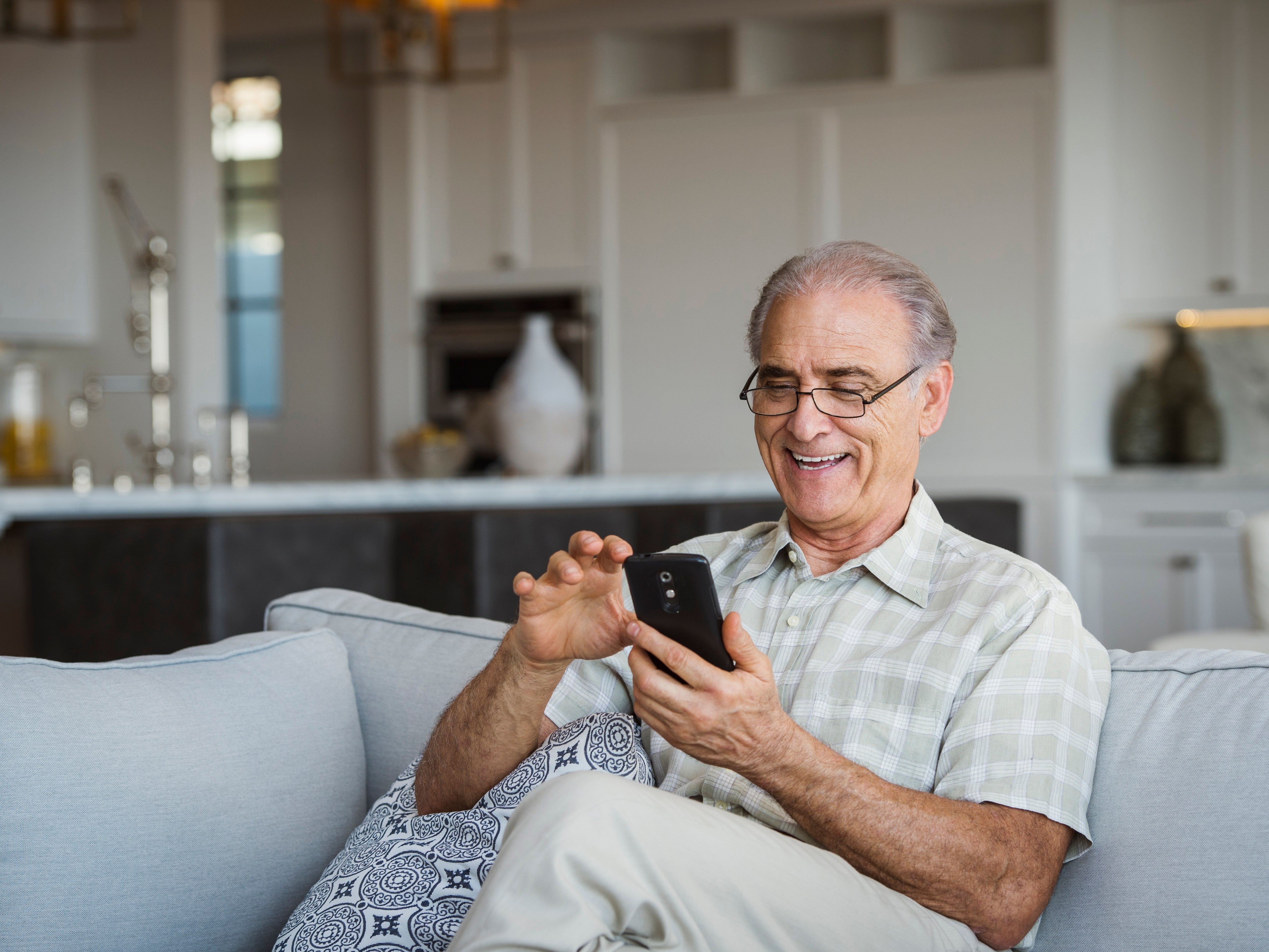 Elderly antheral   smiling, utilizing a smartphone portion    sitting connected  a sofa  successful  a modern   surviving  room
