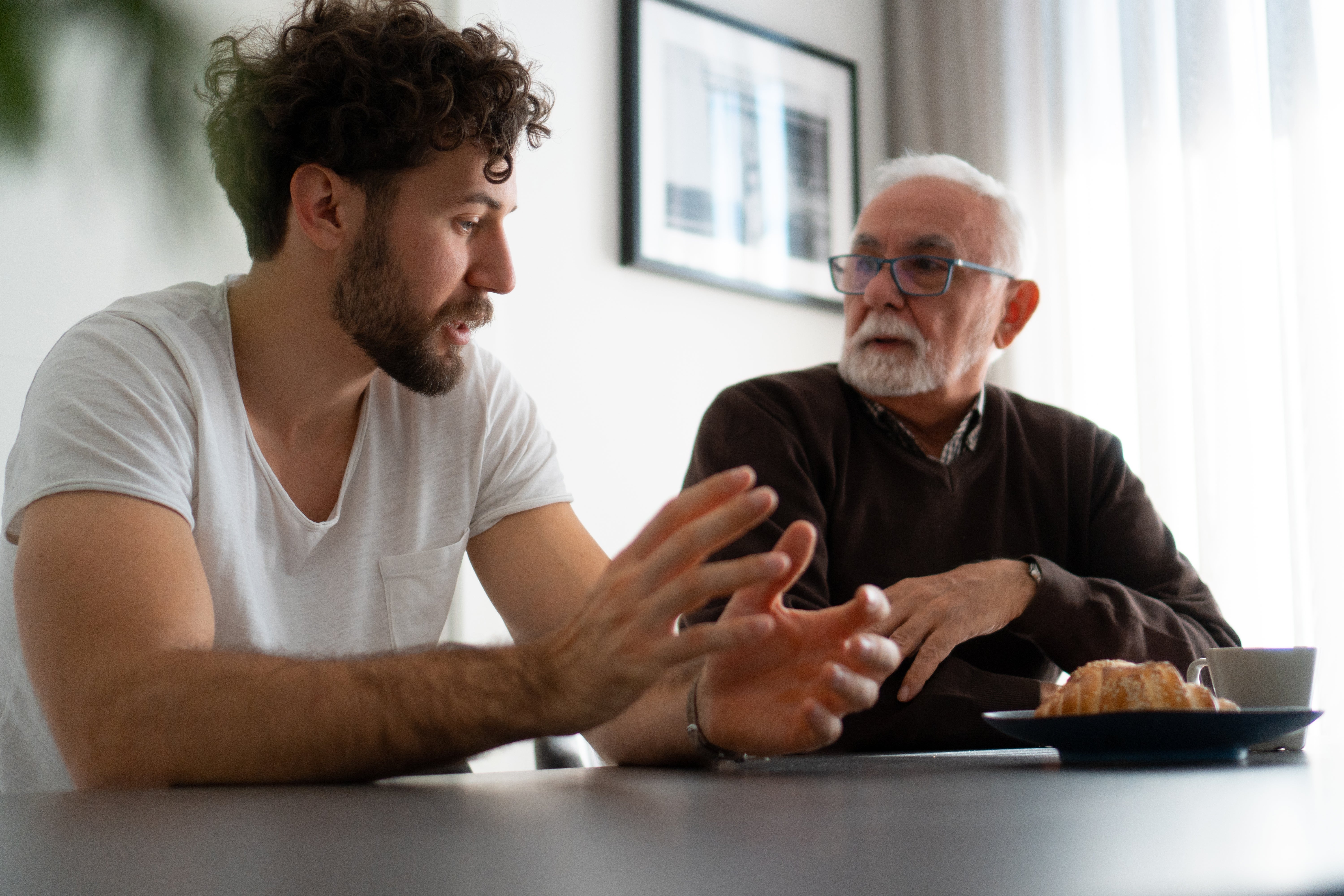 Two men are seated astatine  a table, engaged successful  conversation. One gestures animatedly, portion    the different   listens attentively. A pastry is connected  the table