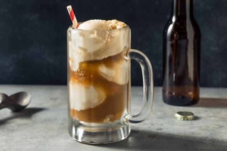 Root beer float with ice cream in a glass mug, topped with a striped straw, on a table next to an empty bottle and bottle cap