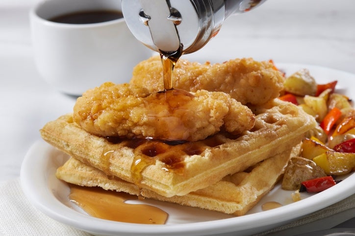 Chicken and waffles on a plate with syrup being poured, accompanied by mixed roasted vegetables and a cup of coffee in the background