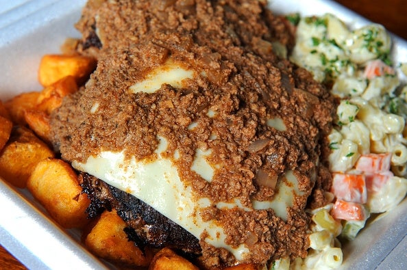 A takeout container holds a juicy beef patty topped with creamy gravy alongside crispy potatoes and a side of macaroni salad with vegetables