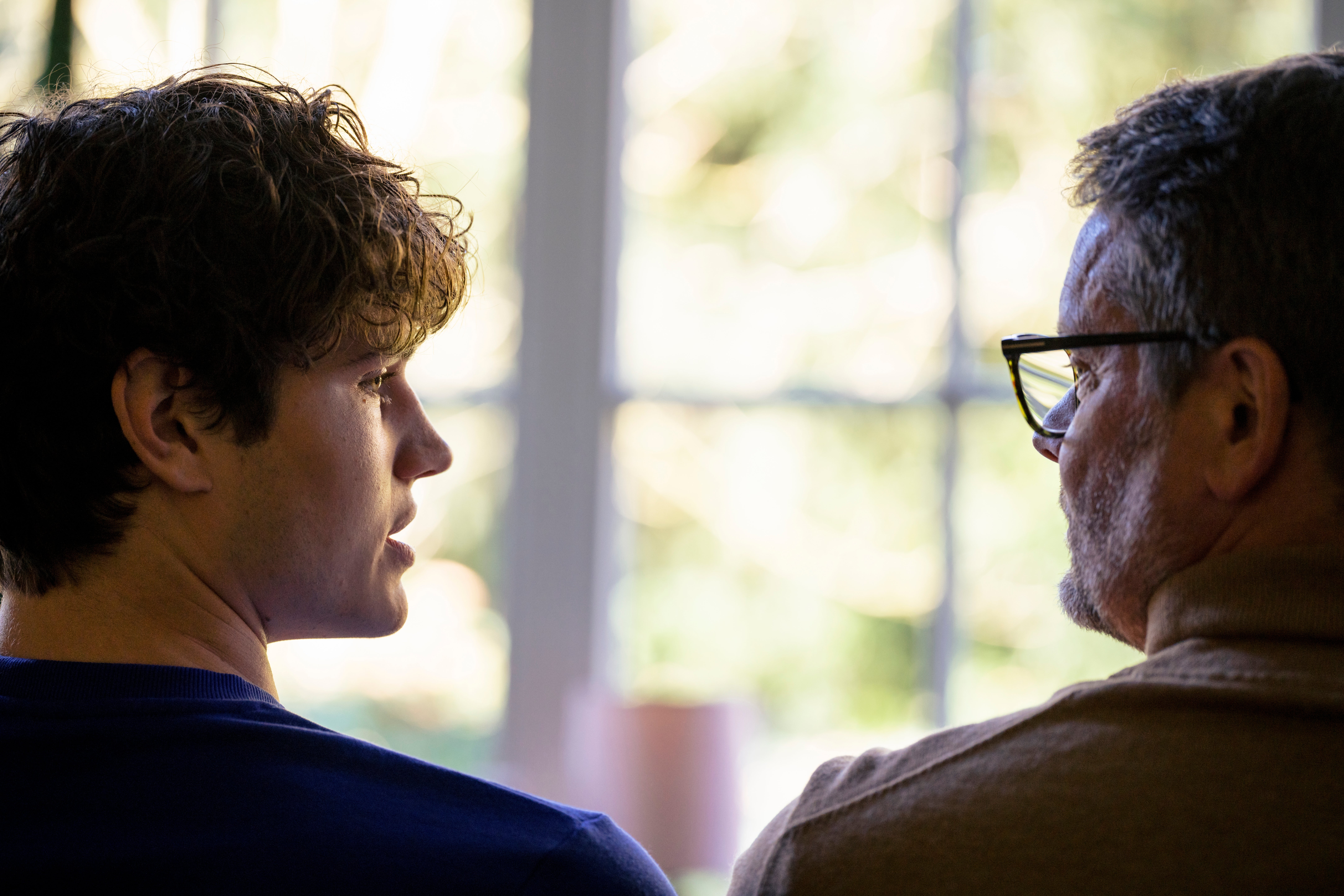 Two radical   sitting and talking, viewed from the side. One wears glasses, and the different   has curly hair. They look  to beryllium  having a superior   conversation