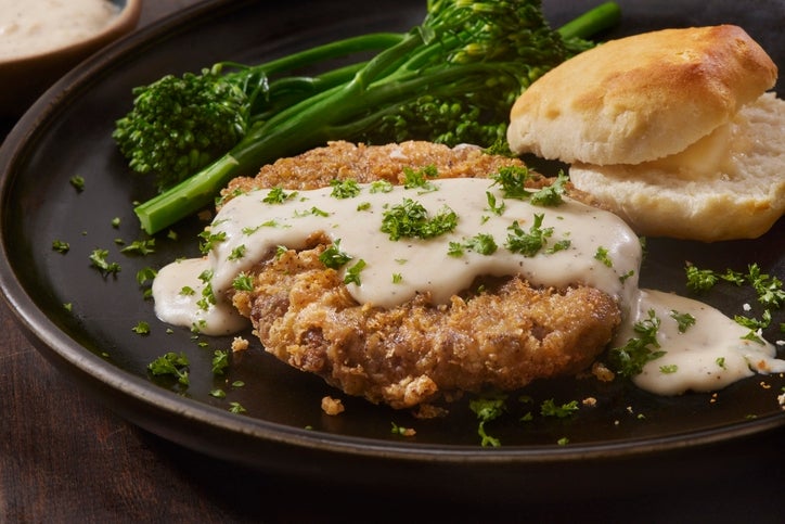 A plate with fried chicken cutlet topped with white gravy, garnished with parsley, served with broccolini and a biscuit