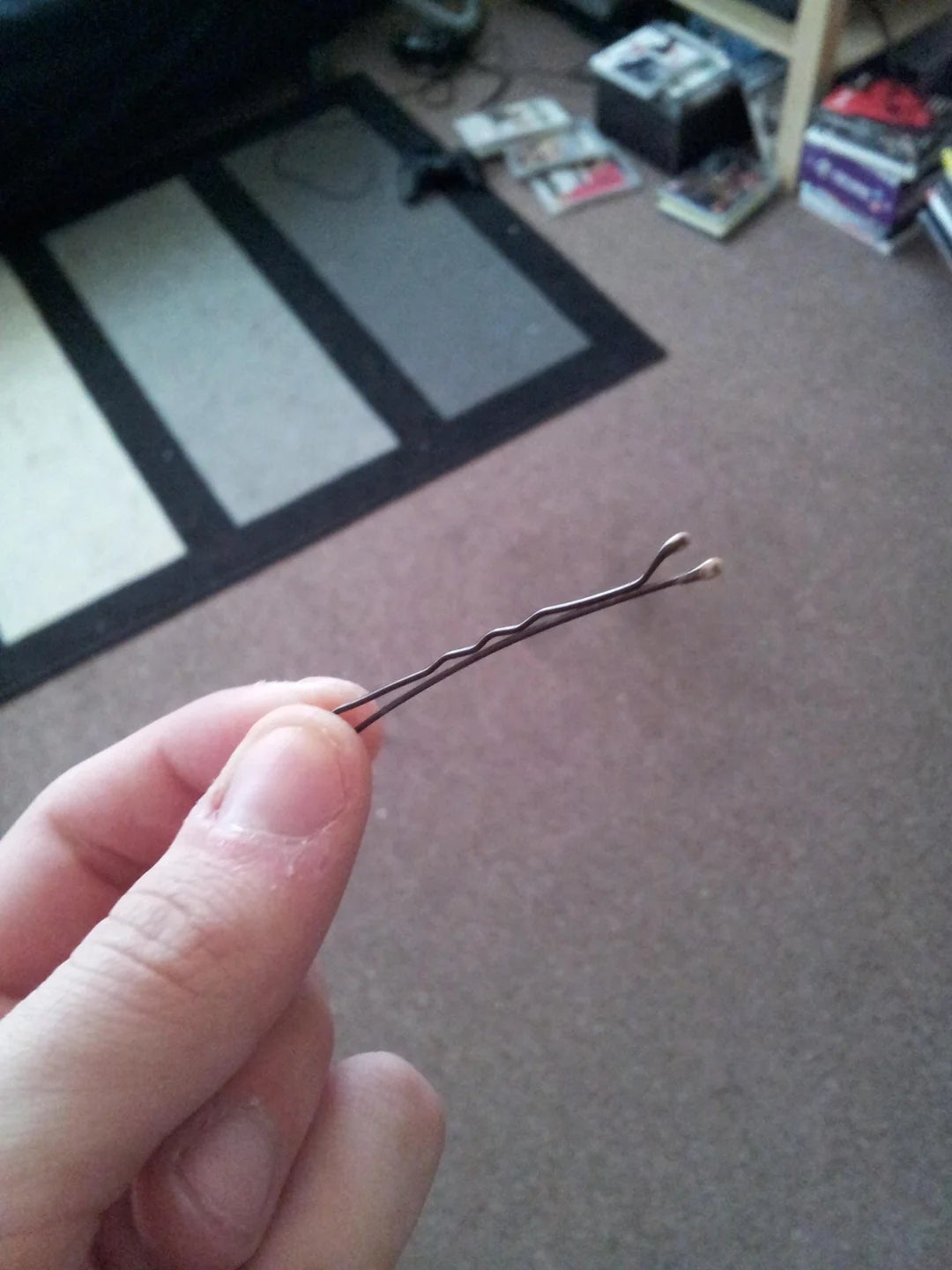 A hand holds a bobby pin. In the background, there are games and a controller on the floor