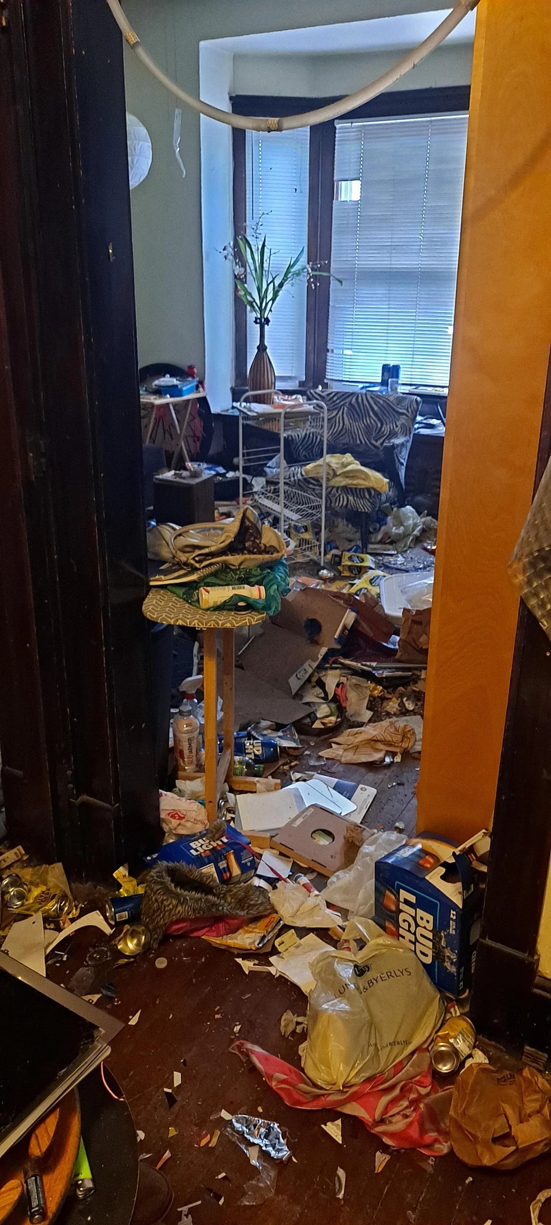 Cluttered room with debris on the floor, including boxes, papers, and trash. A small table holds a vase with flowers. Minimal natural light