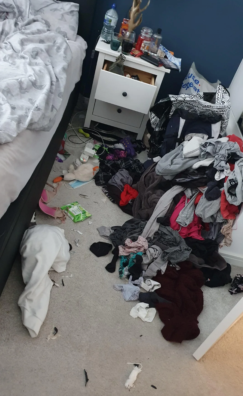 Messy bedroom with unmade bed, cluttered nightstand, and pile of clothes on the floor. Various items scattered, including bottles and packaging