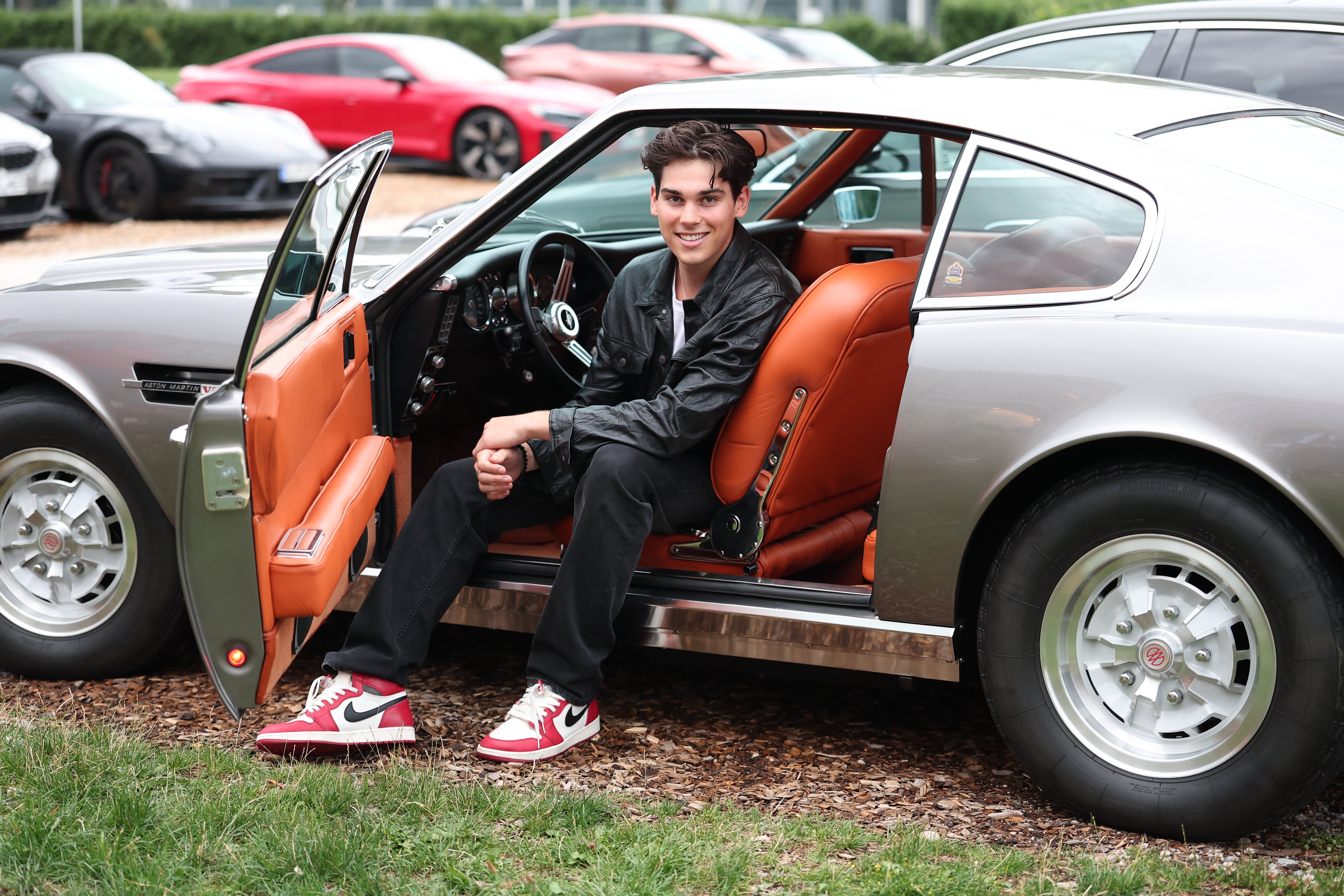 A idiosyncratic   sitting and smiling successful  the unfastened  doorway  of a classical  car, wearing a stylish overgarment   and sneakers. Several modern   cars disposable   successful  the background
