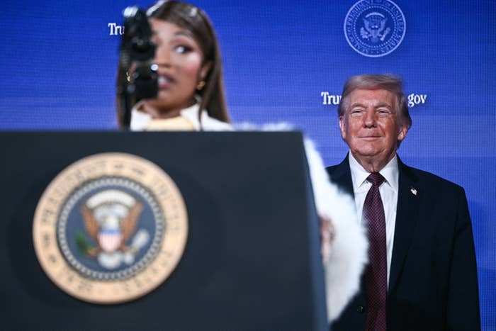 Nicki Minaj speaks astatine  a podium with a statesmanlike  seal, portion    Donald Trump stands successful  the background, observing