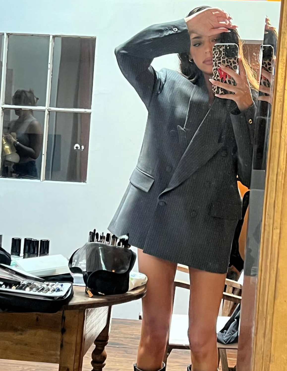 Person taking a reflector  selfie, wearing an oversized blazer and holding a telephone  with a leopard people     case. Makeup items are disposable   connected  a array  nearby