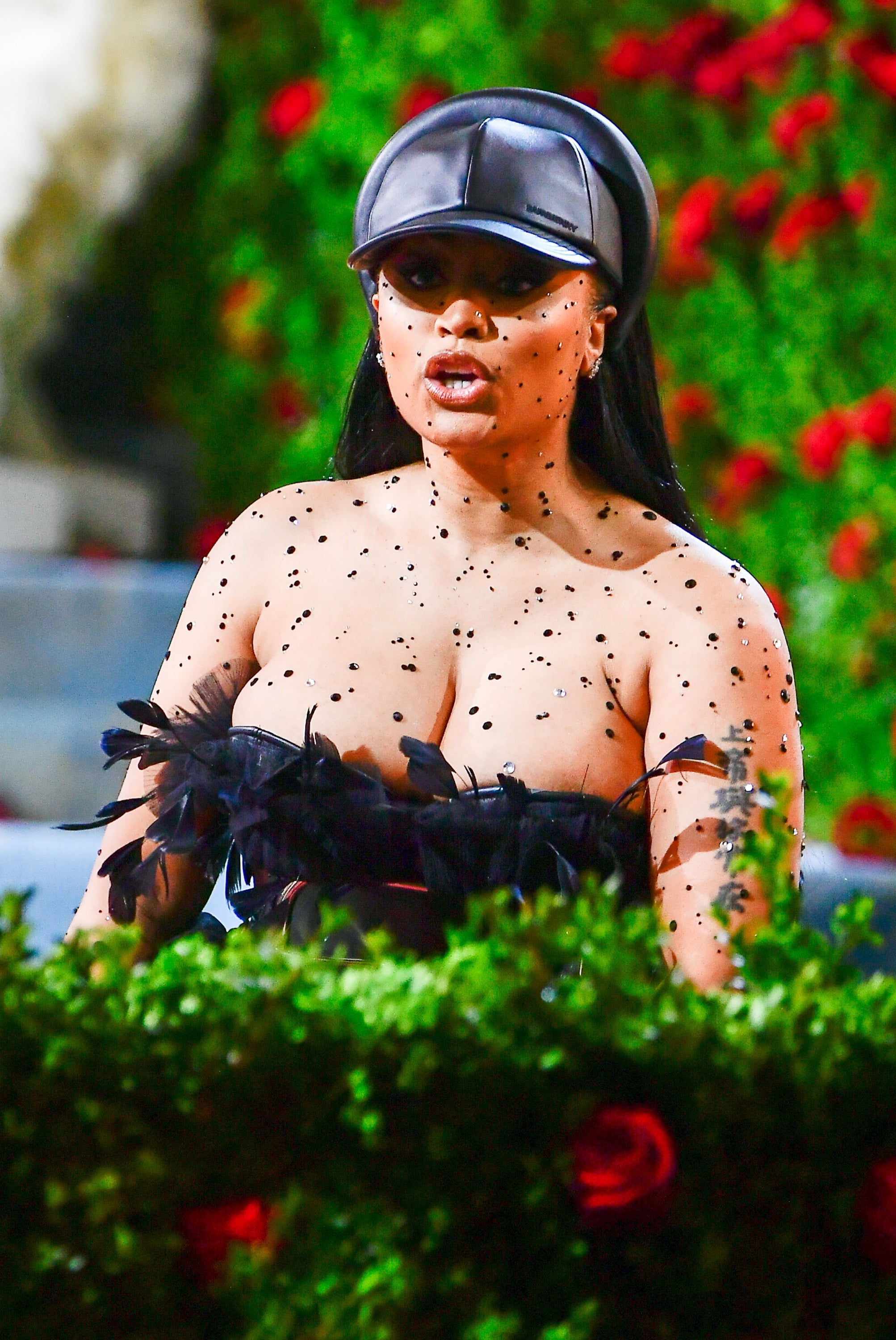 Nicki Minaj successful a strapless formal adorned with feathers and polka dots, wearing a visor cap, posing connected a floral-themed reddish carpet