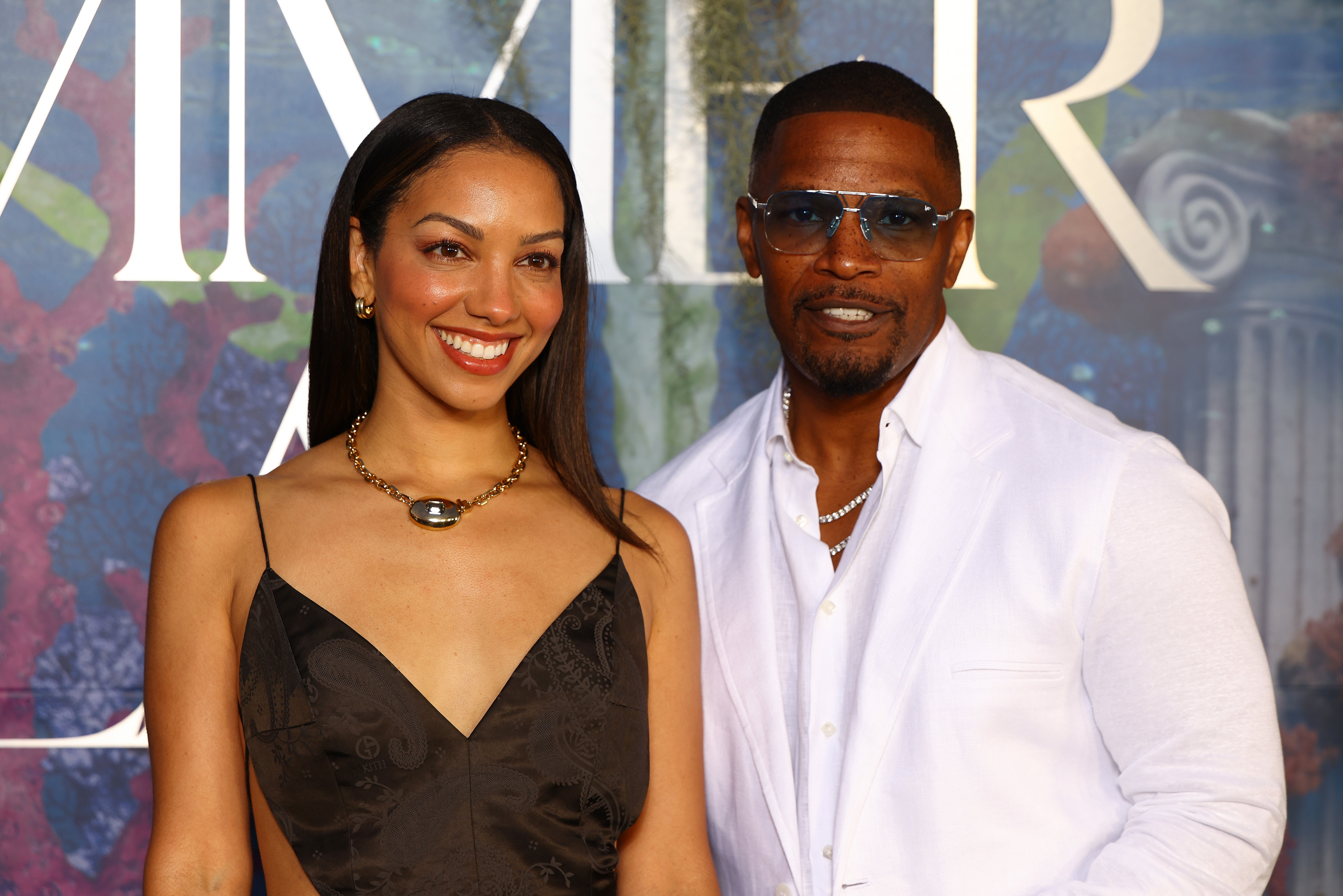 Two radical   airs  connected  a reddish  carpet; 1  wears a sleeveless V-neck dress, the different   a achromatic  garment  and sunglasses