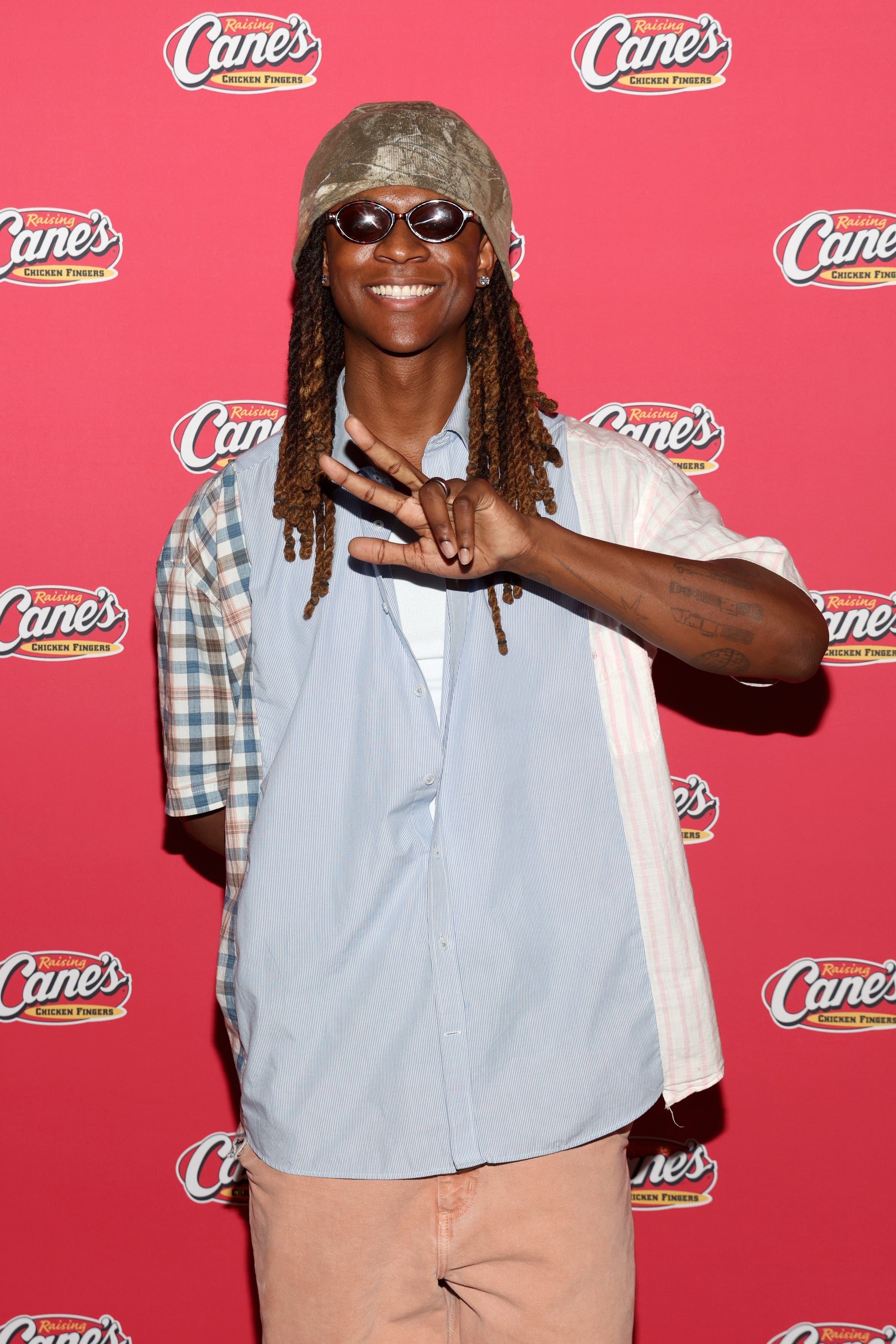 Person posing successful  mixed-pattern garment  with sunglasses and hat, making a bid    motion   connected  a reddish  carpet with Raising Cane's logos successful  the background