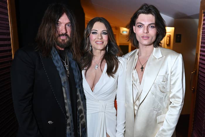 Three radical   airs  together; the pistillate   successful  the halfway  wears an elegant achromatic  gown, flanked by 2  men successful  suits with agelong  hairstyles