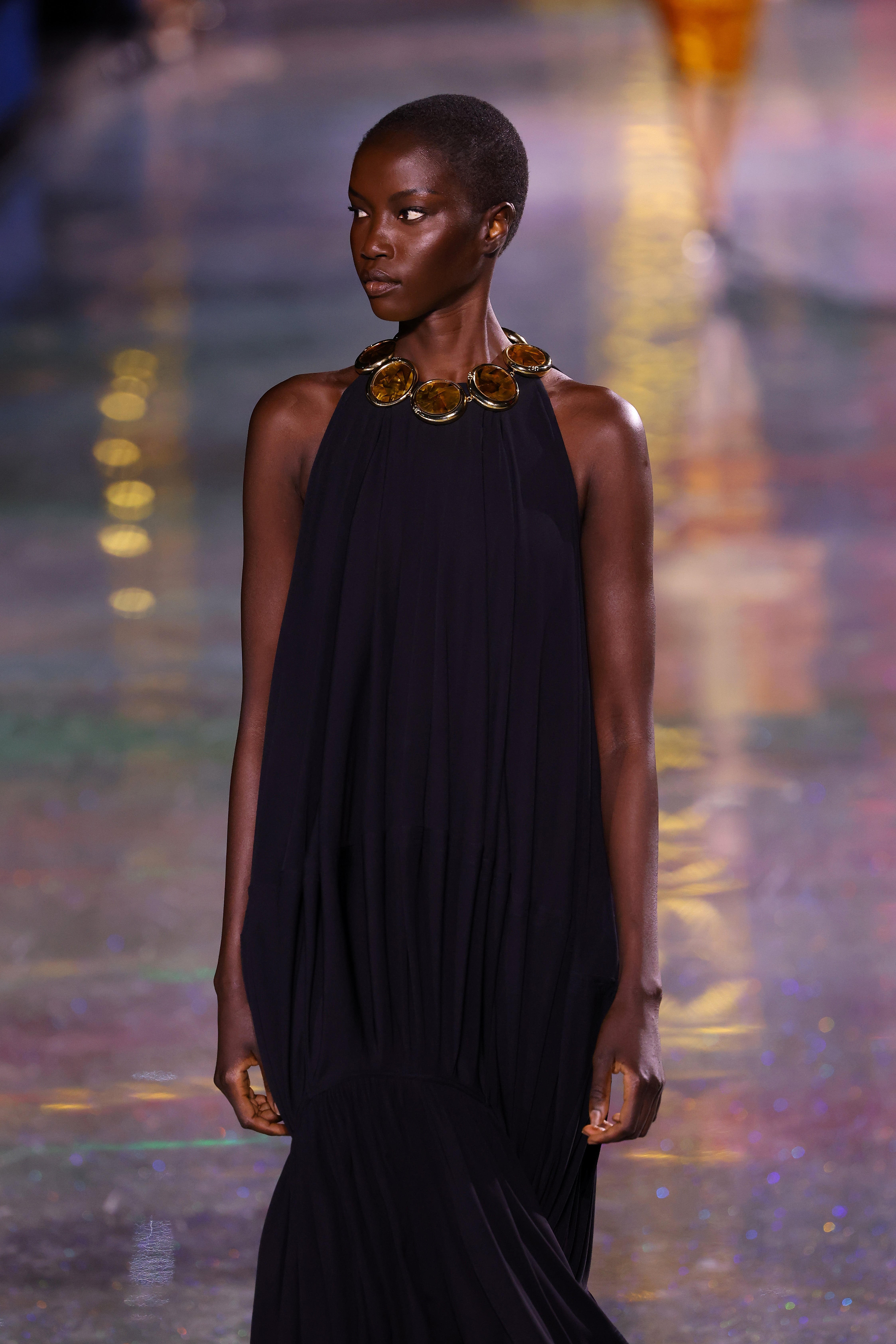 A exemplary  walks the runway successful  a sleeveless, flowing acheronian  gown with a distinctive neckpiece during a manner  show
