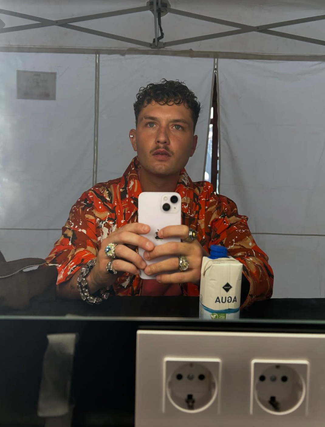 Person takes a reflector  selfie, wearing a patterned garment  and holding a phone. A portion  tin  and plugs are disposable   connected  the counter