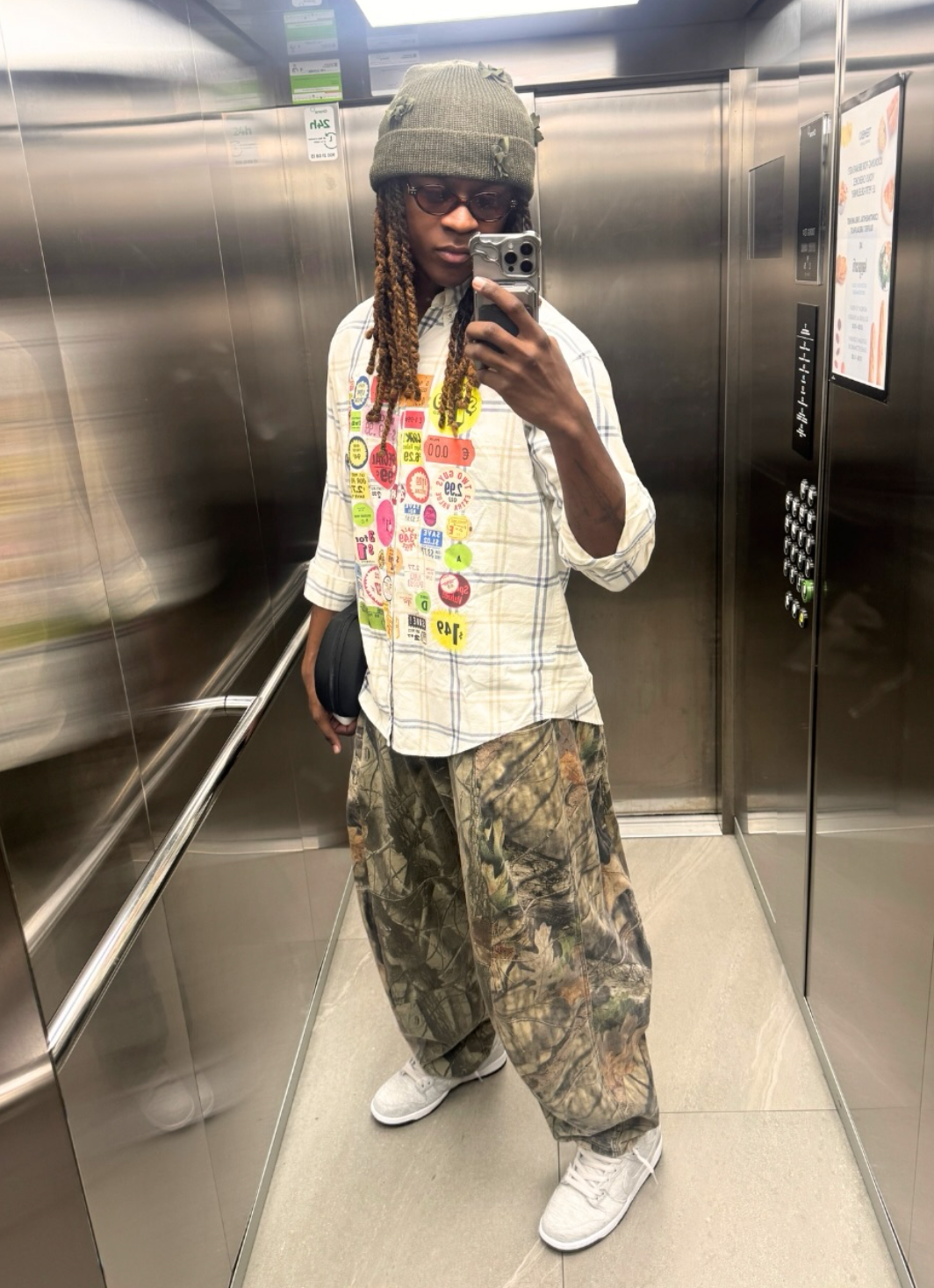 Person successful  an elevator taking a selfie, wearing a checkered garment  with colorful patches, camo pants, sneakers, sunglasses, and a knitted beanie