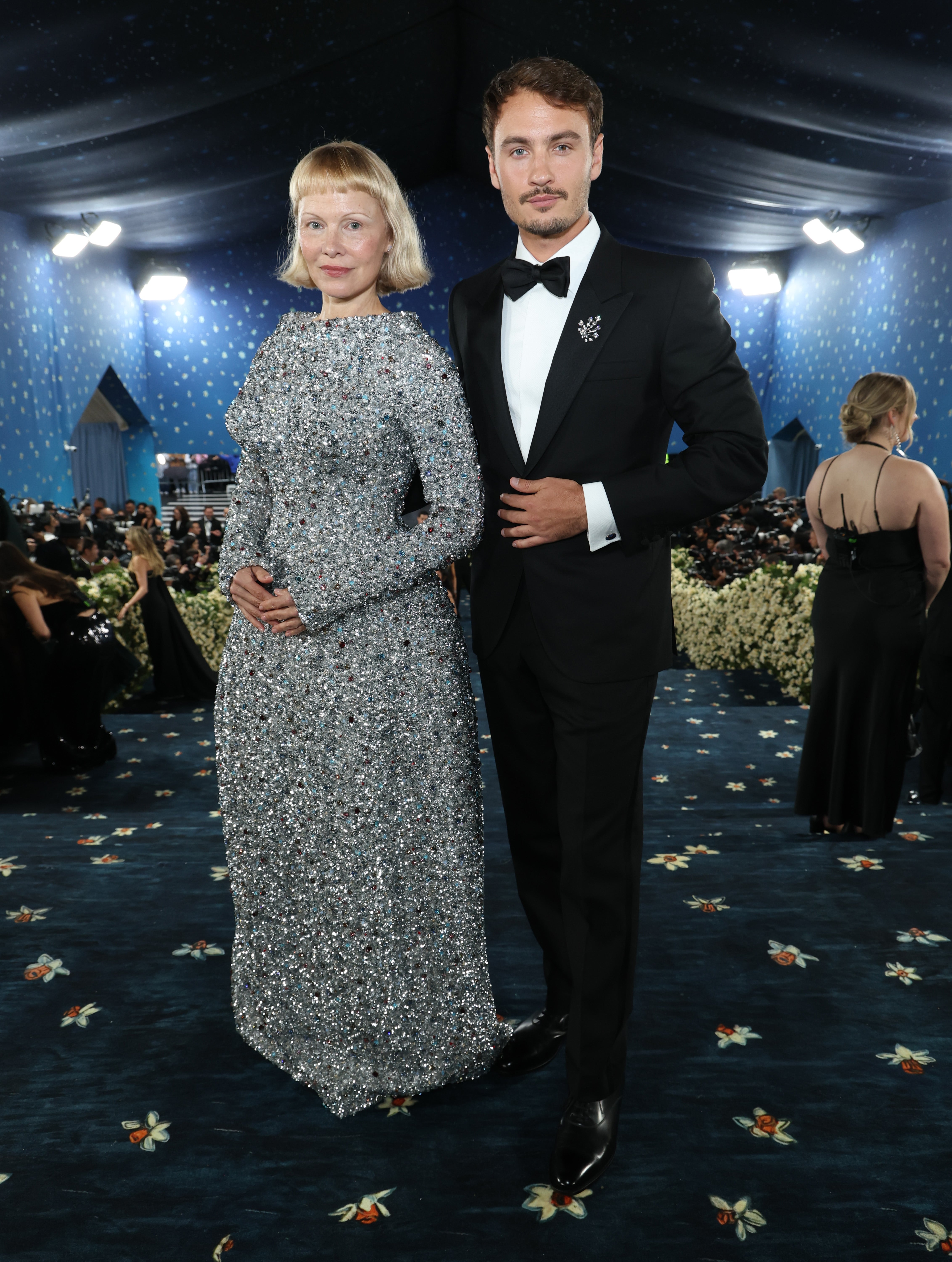 Two radical   connected  a ceremonial  reddish  carpet. The idiosyncratic   connected  the near  wears a sparkling agelong  dress. The idiosyncratic   connected  the close    wears a tuxedo with a bow tie