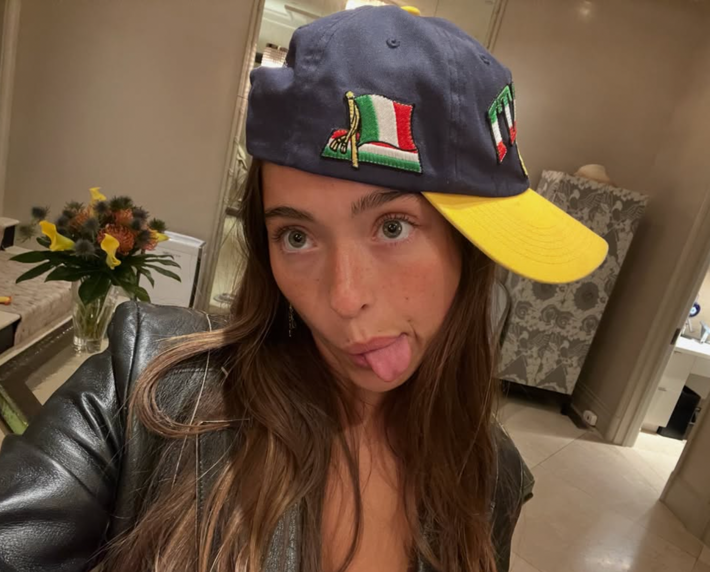 Person playfully sticking retired  tongue, wearing a sideways headdress  with embroidered plan  and a casual leather overgarment   indoors