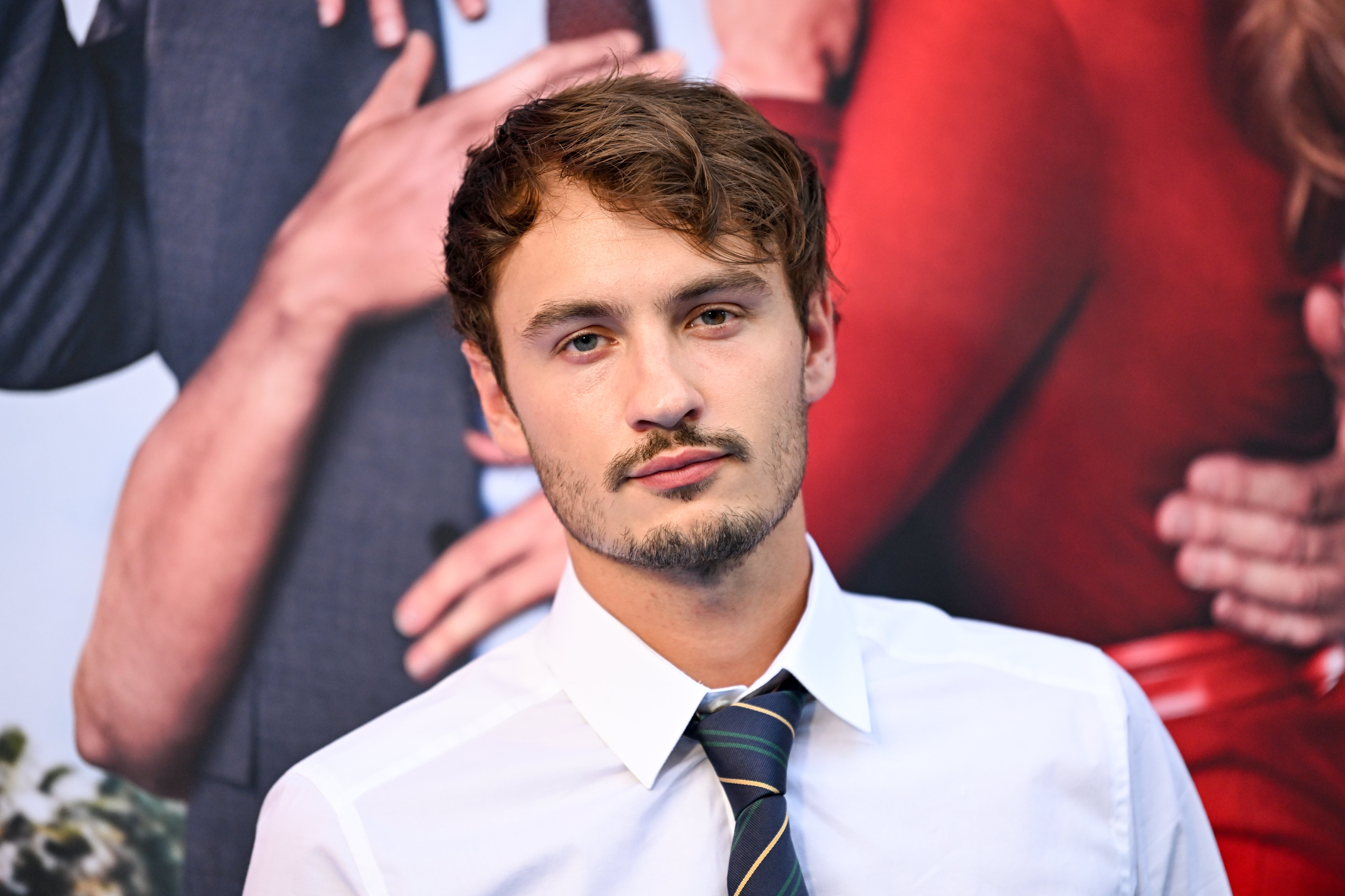 A idiosyncratic   poses connected  a reddish  carpet successful  a achromatic  garment  and striped tie, with a blurred inheritance  of different   people's cropped hands and outfits