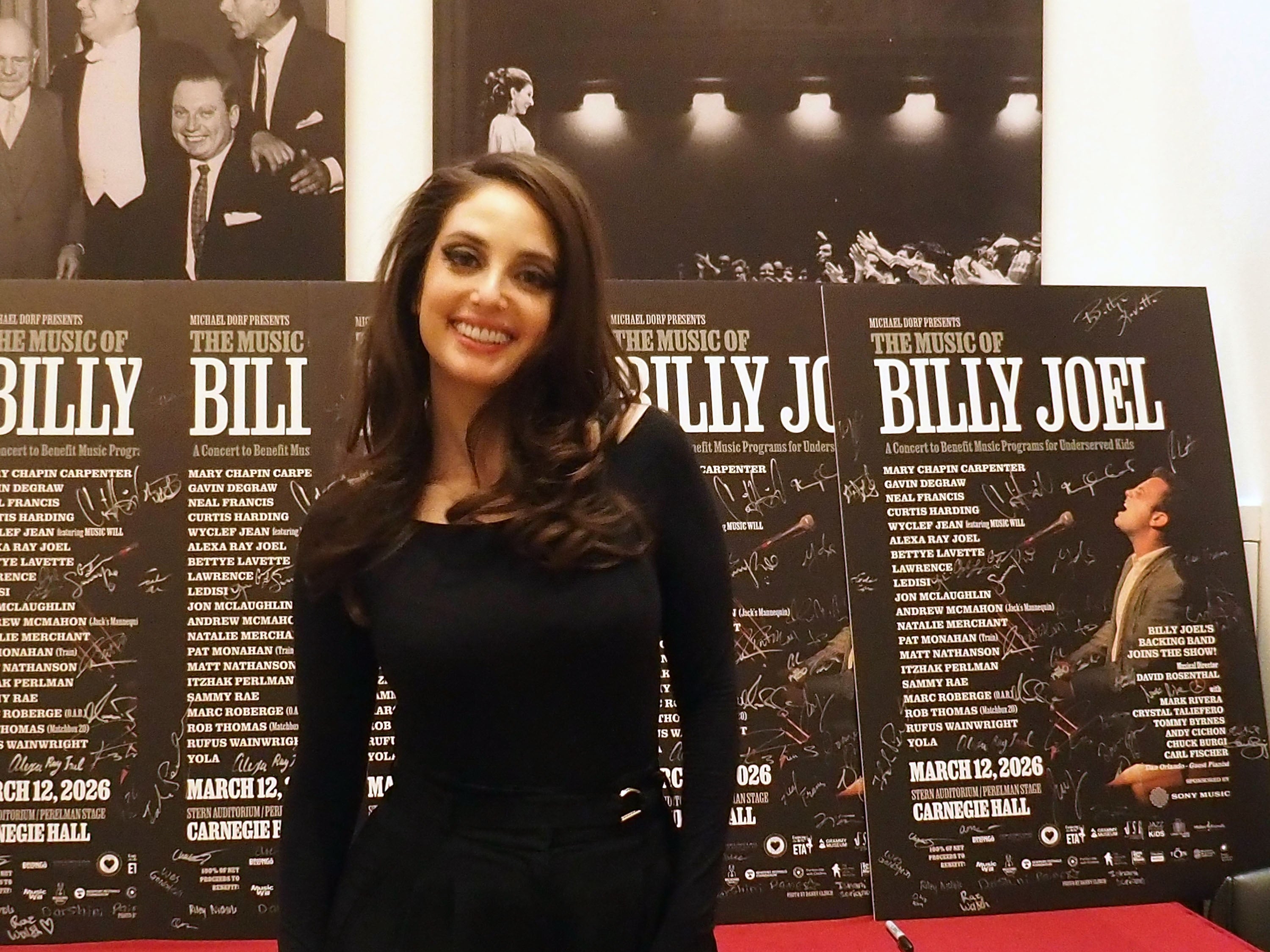 A idiosyncratic   poses astatine  an lawsuit   successful  beforehand   of posters for "The Music of Billy Joel" concert. They are wearing elegant, modern   attire