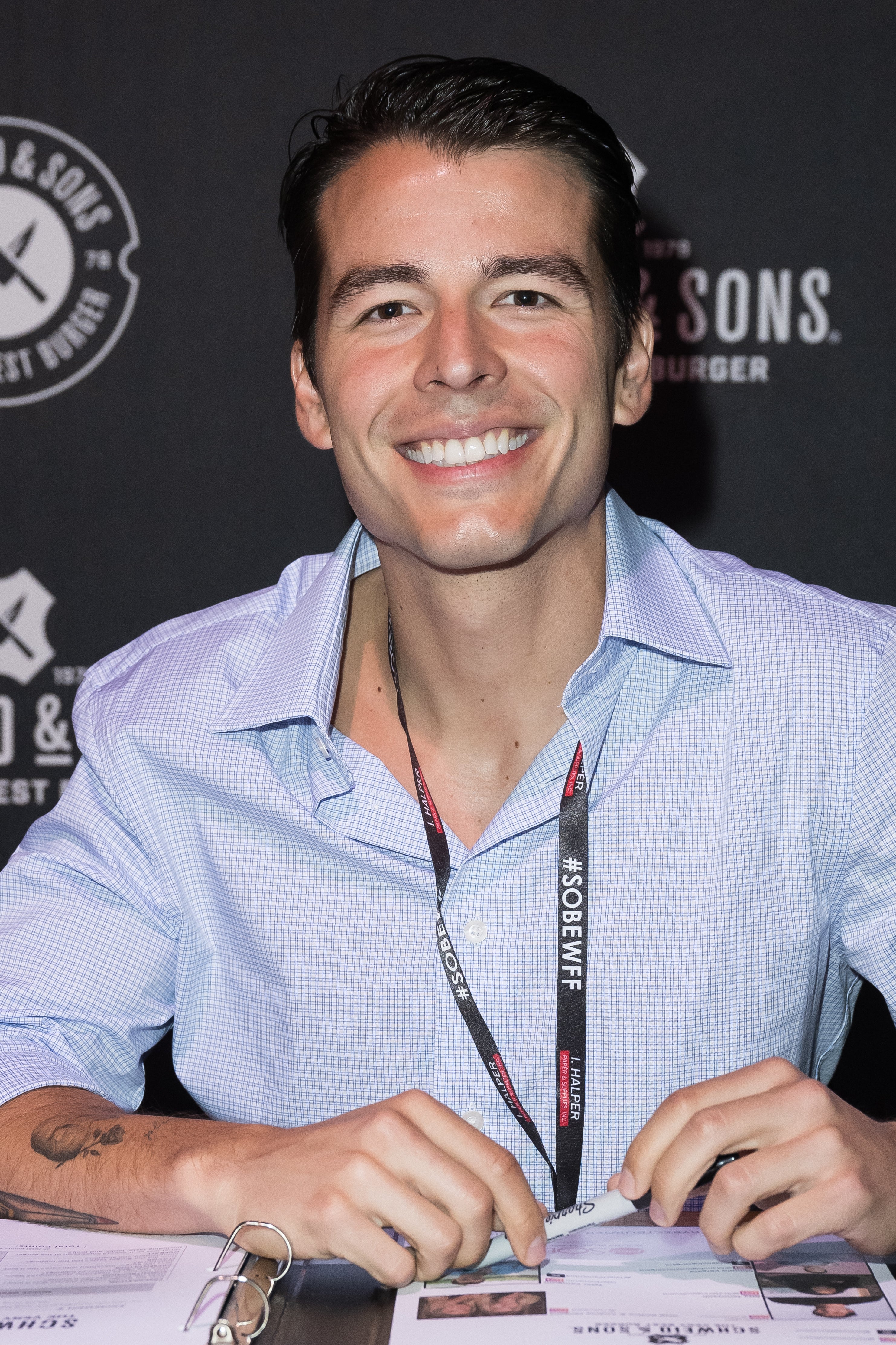 Person smiling, wearing a casual button-up shirt, seated astatine  an lawsuit   array  with a booklet and lanyard visible