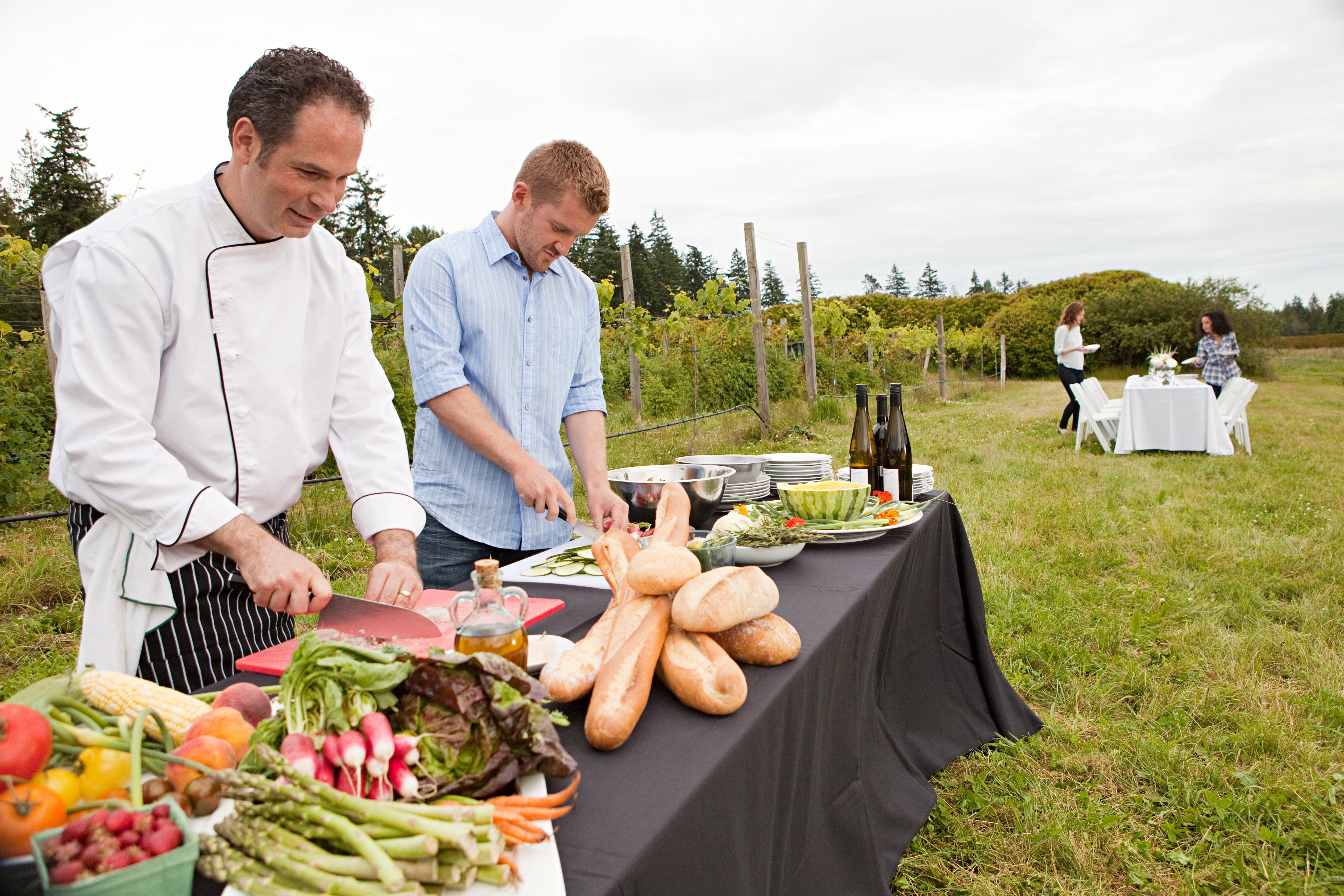 Chefs hole nutrient astatine an outdoor array with assorted vegetables, bread, and vino bottles; a mates dines astatine a array successful the background