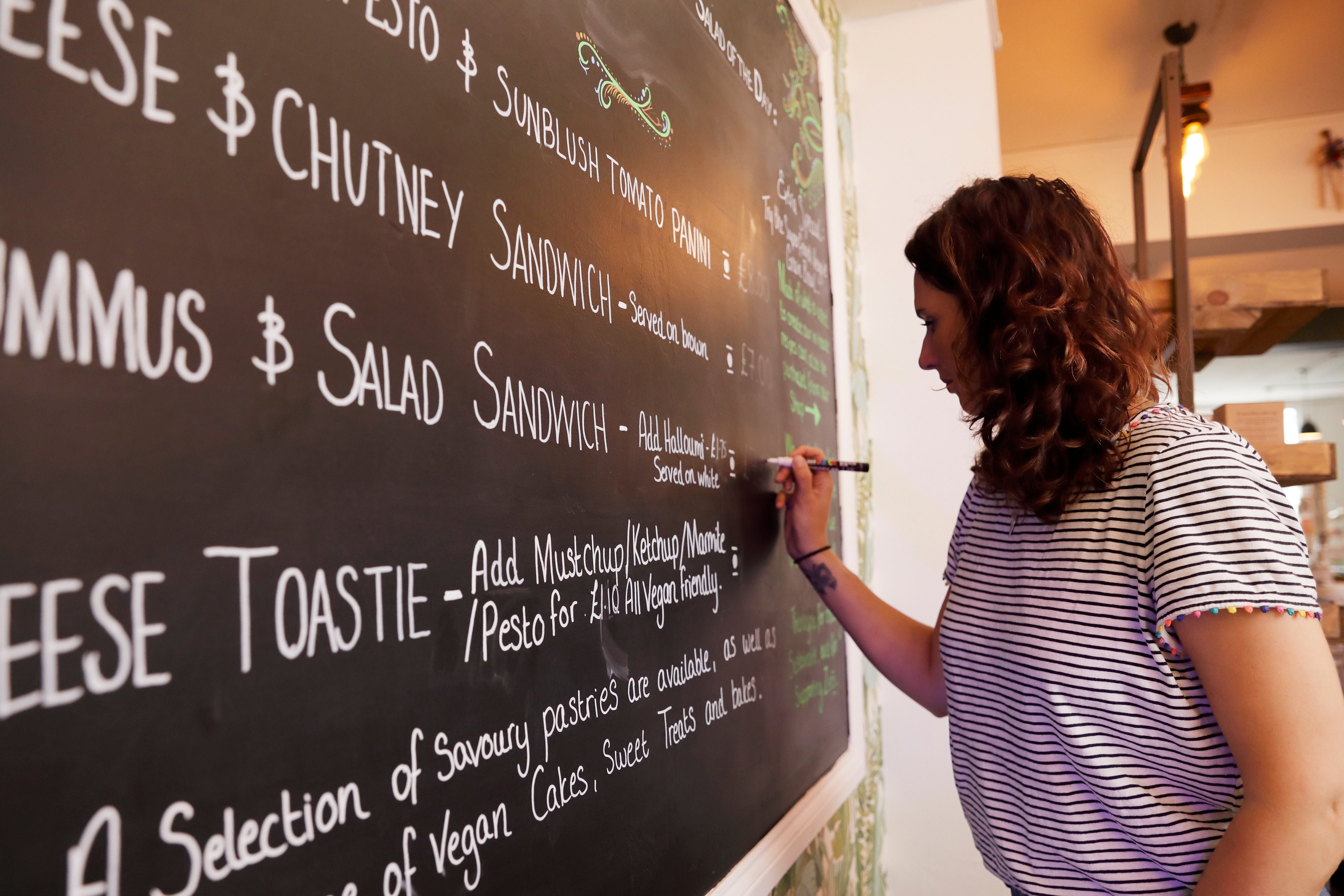 Person penning paper items connected a chalkboard successful a restaurant. Menu includes sandwiches, hummus, salad, and food toastie options