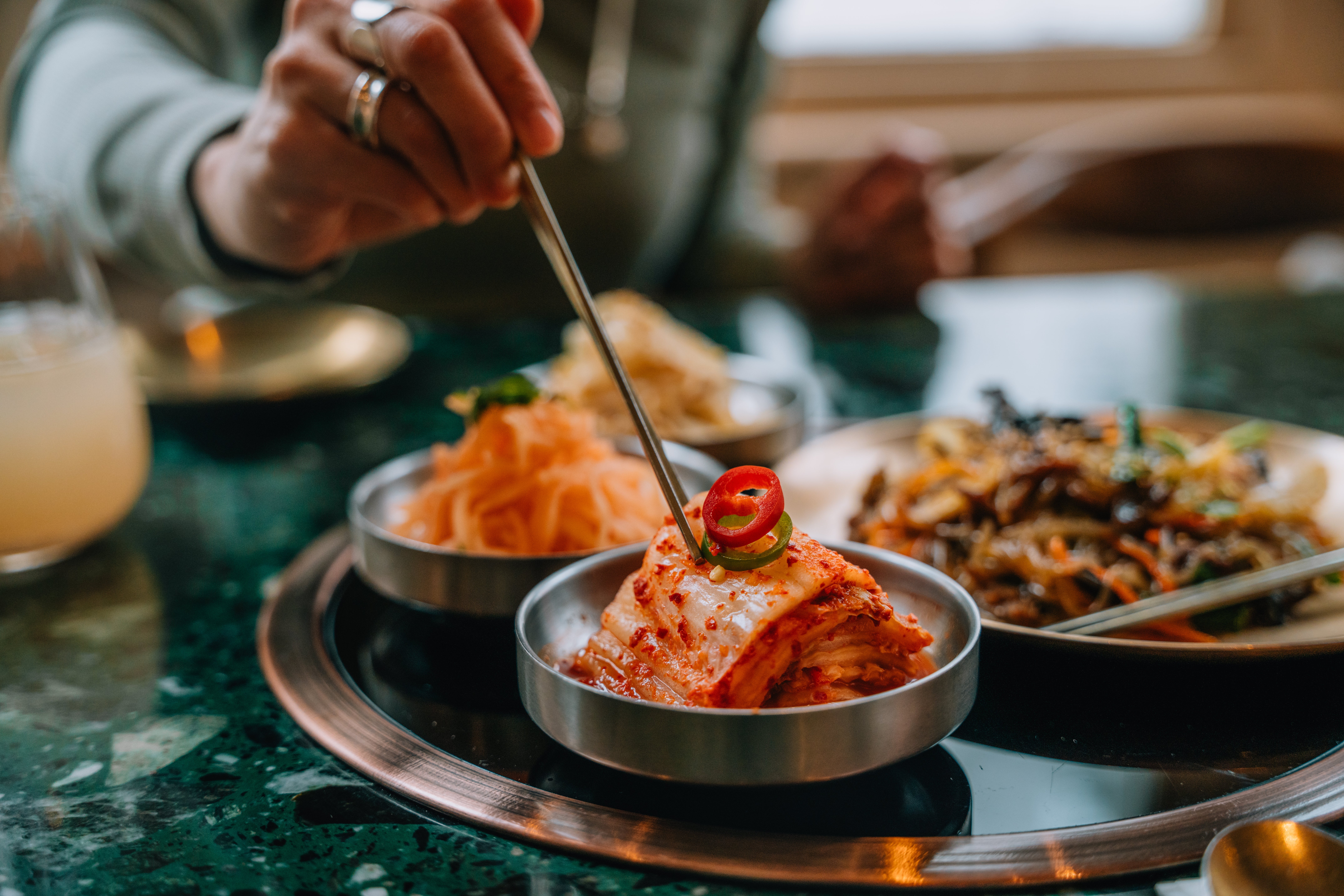 Person serving kimchi and broadside dishes from a tray with chopsticks, featuring assorted Korean dishes successful tiny bowls