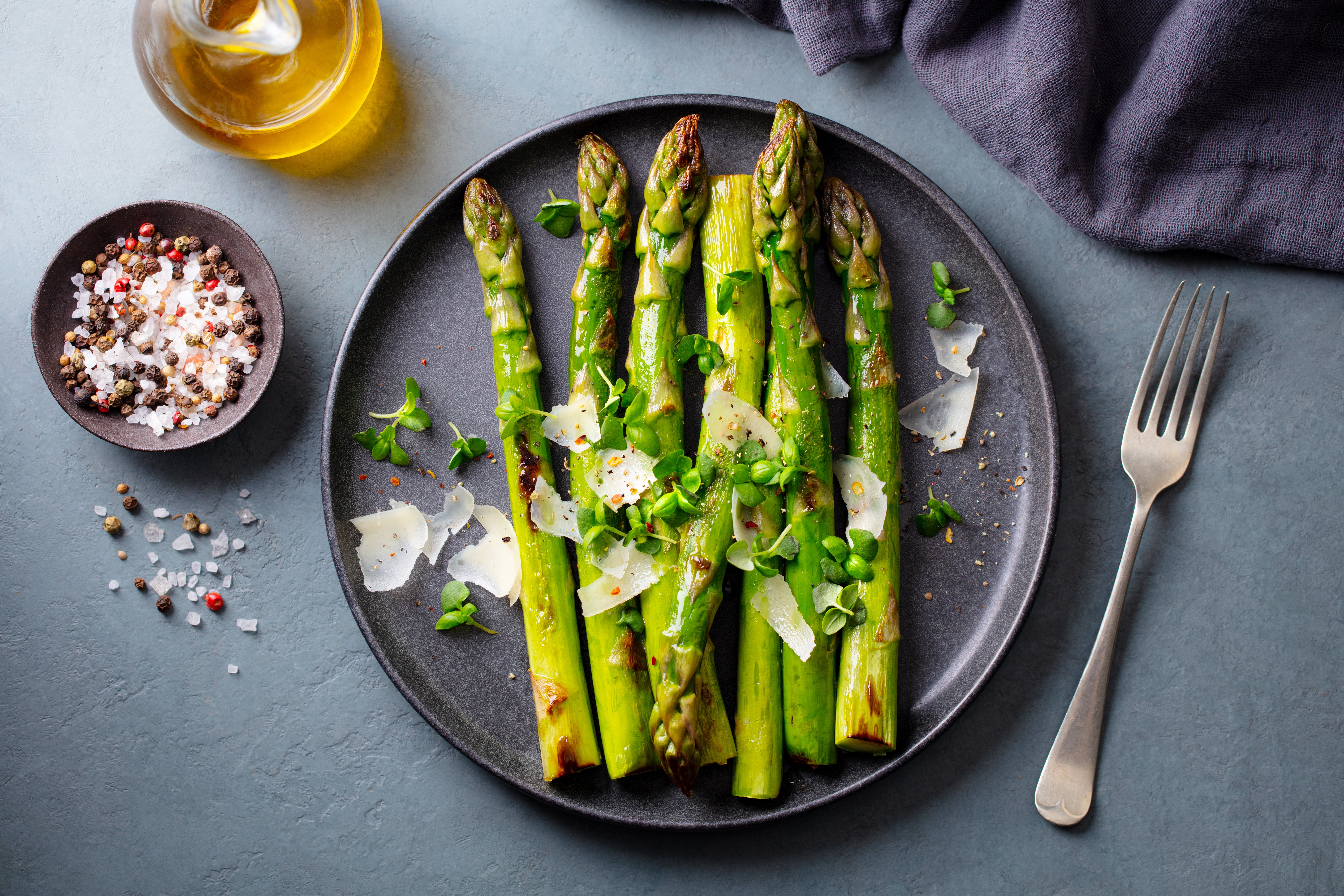 Grilled asparagus connected a plate, garnished with parmesan shavings, herbs, and pepper. Olive lipid and a fork are beside the plate