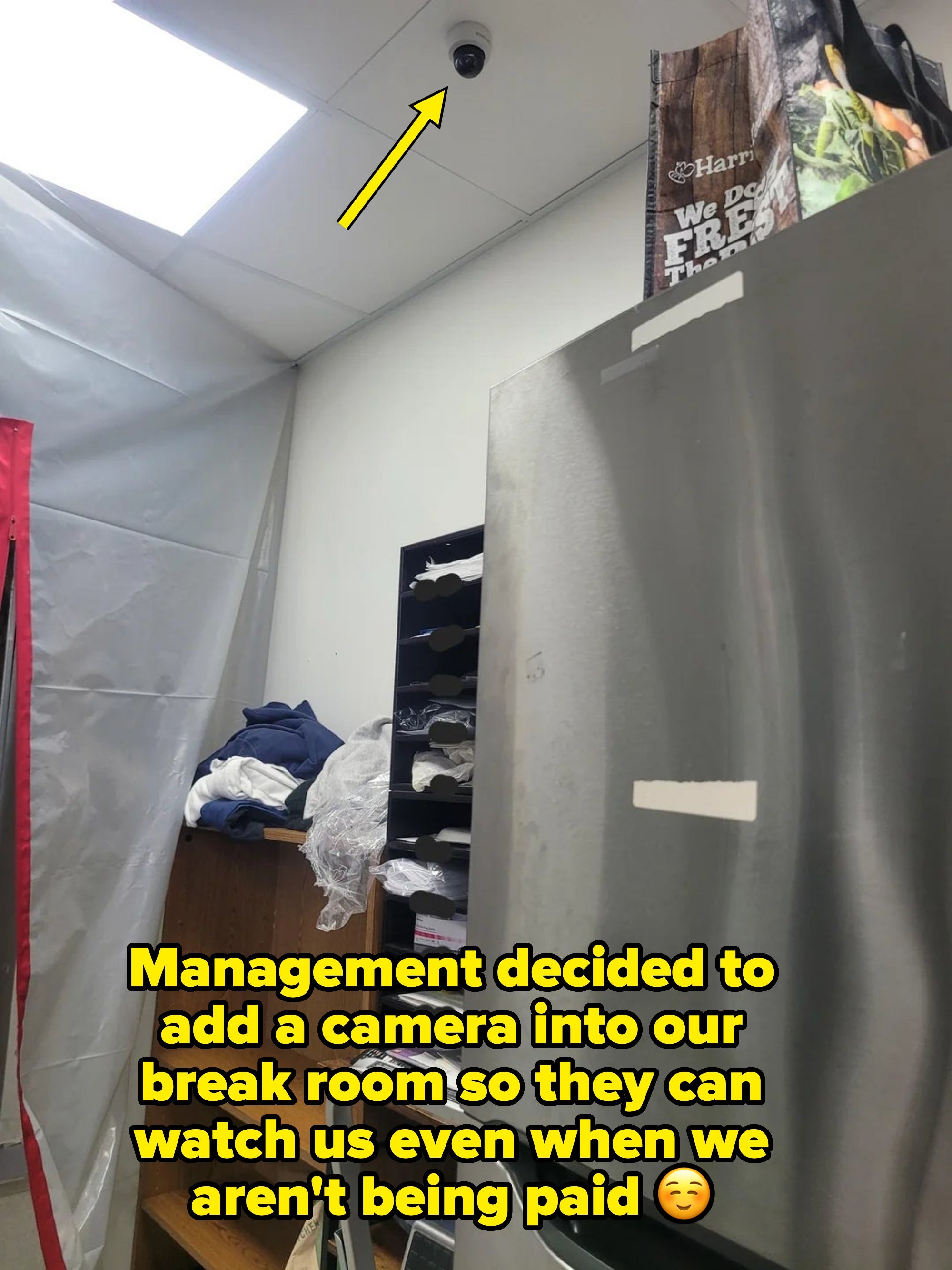 Room with a metal fridge, open shelving, and a stack of bags on top. Ceiling camera visible