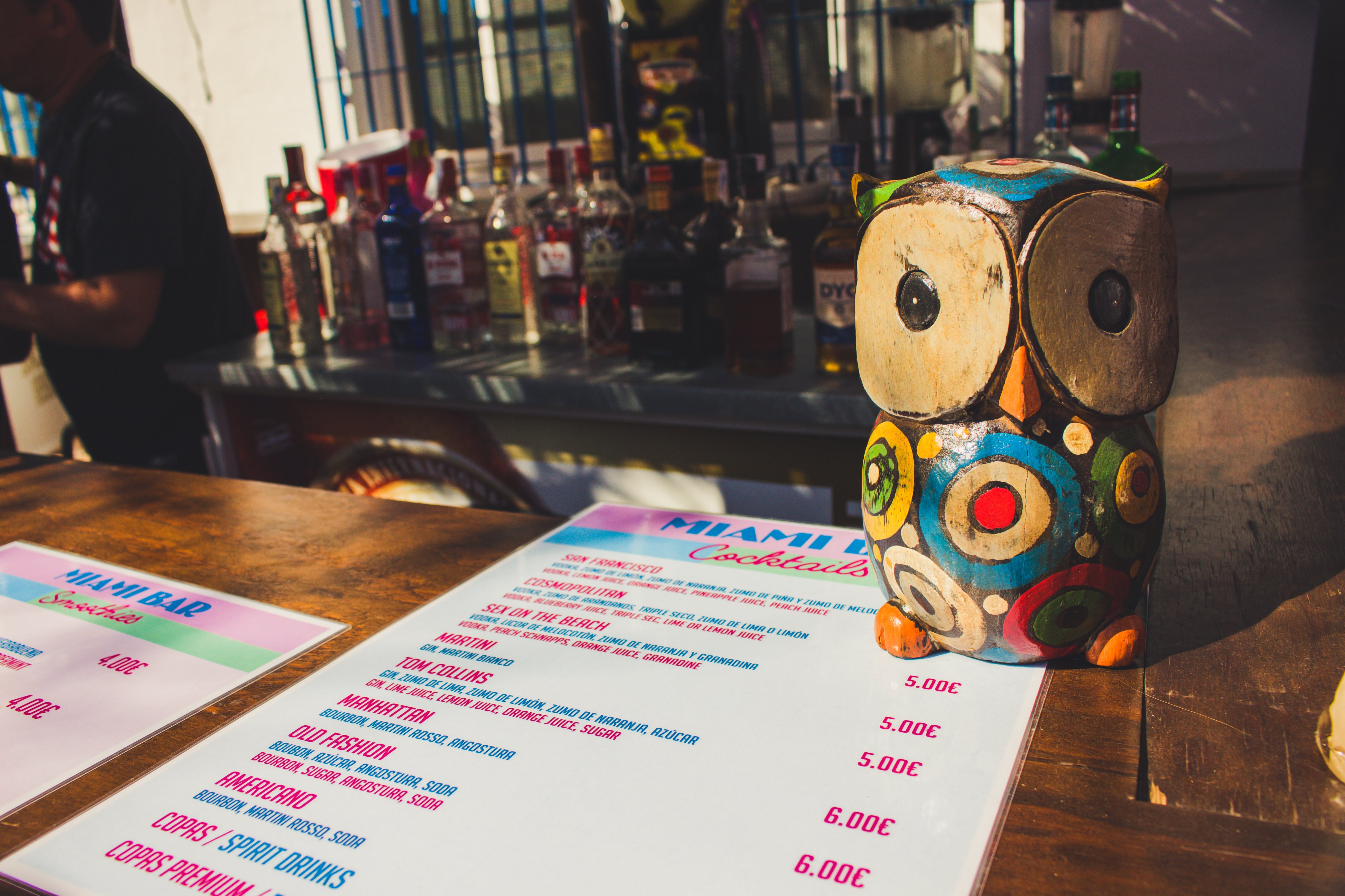 Bar antagonistic with assorted liquor bottles, an owl-shaped decoration, and a drinks paper listing cocktails and prices successful a sunny outdoor setting