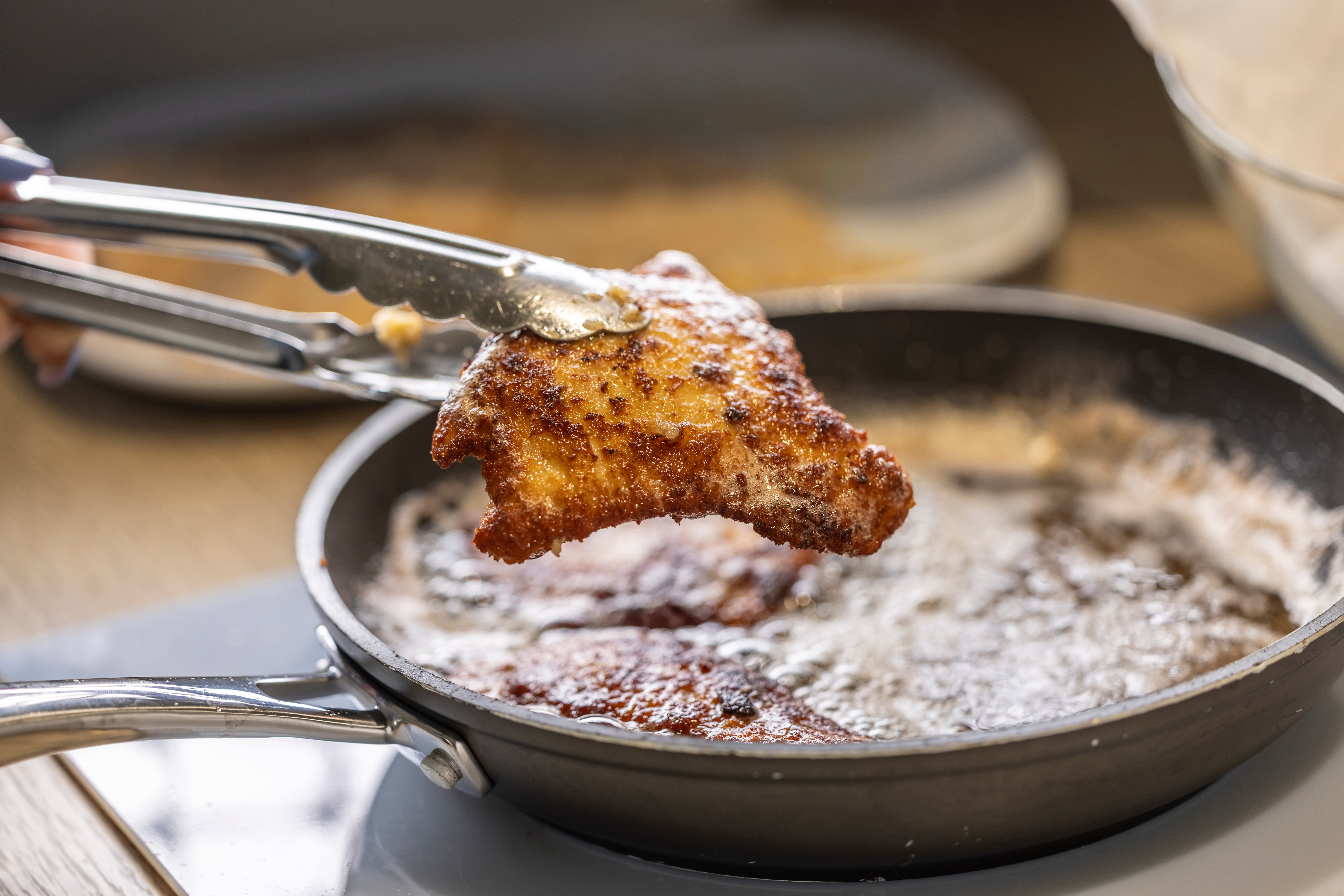 Tongs holding a portion of fried chickenhearted implicit a sizzling pan, highlighting a home-cooking scene