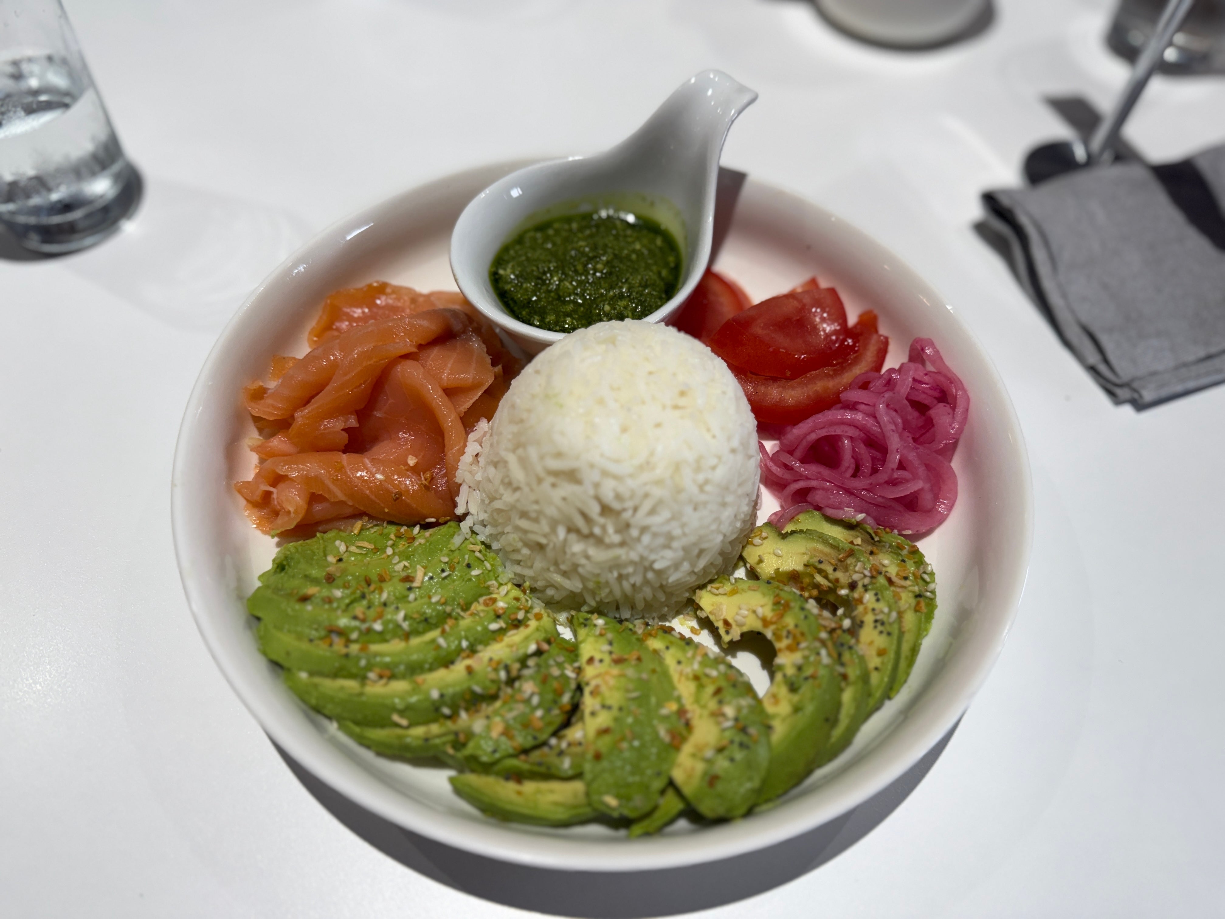 Bowl with avocado slices, smoked salmon, rice, reddish onions, tomatoes, and greenish sauce. Plateware includes a napkin and utensils