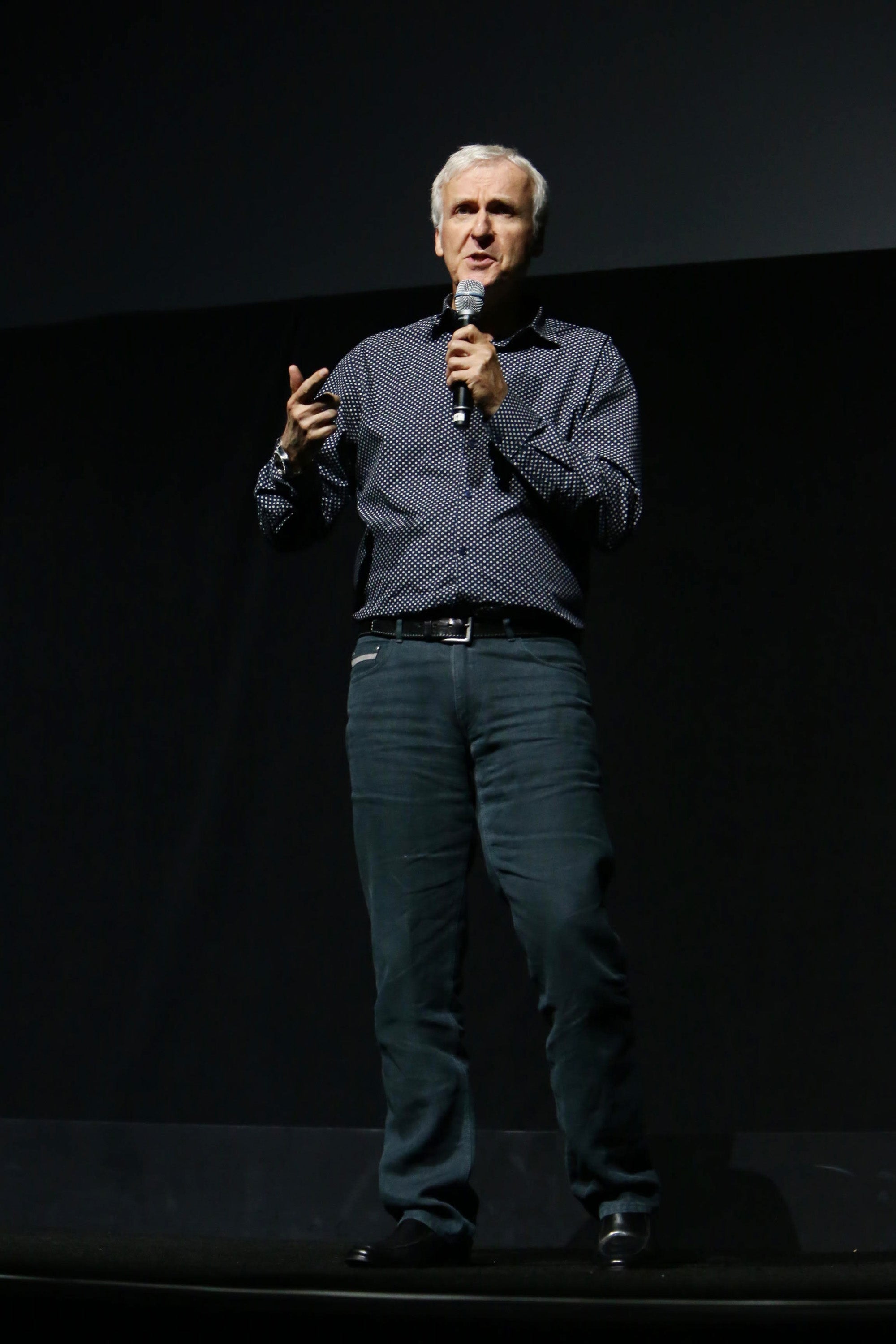 James Cameron in a patterned shirt and jeans speaks on stage, holding a microphone in one hand