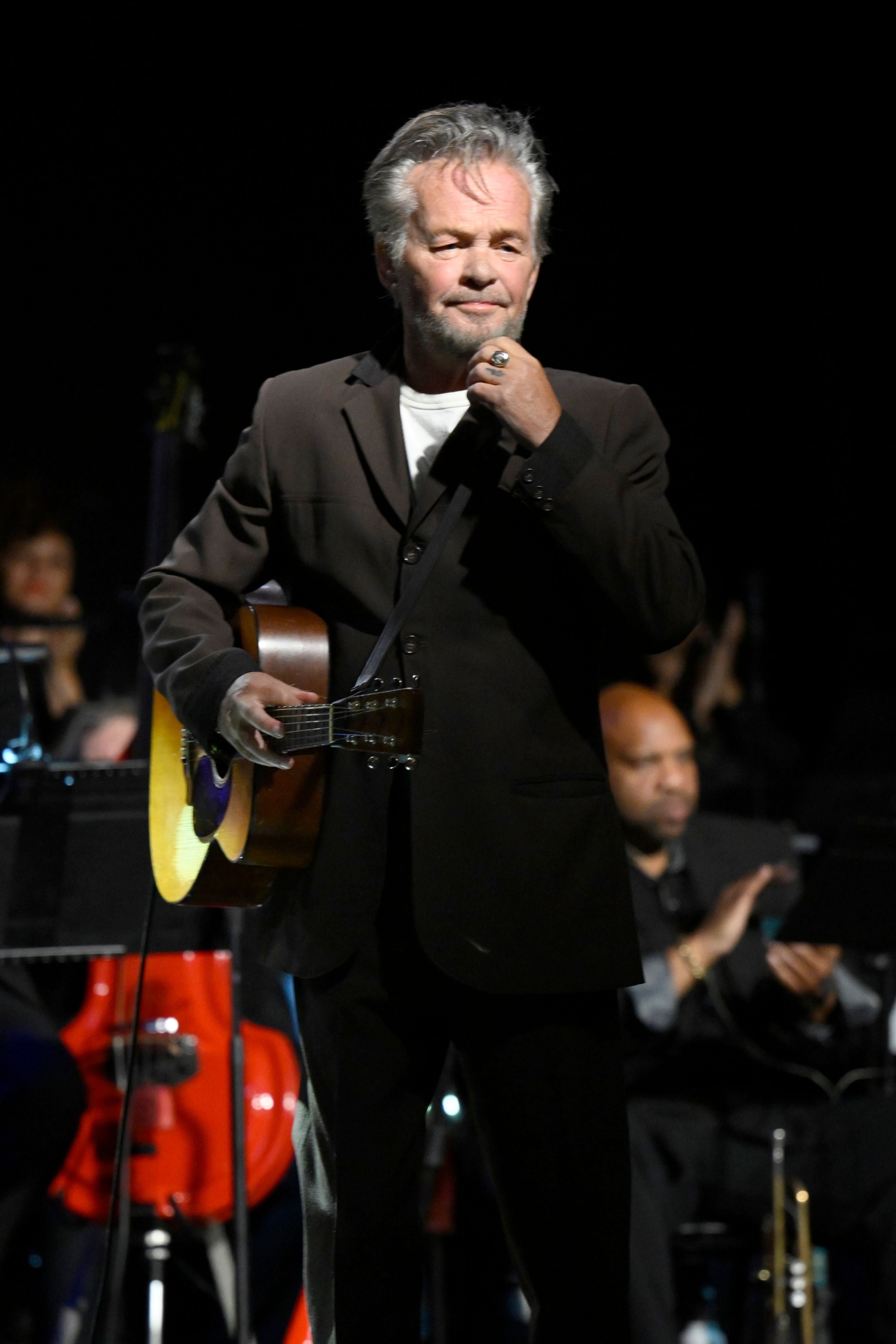 John Mellencamp is on stage holding a guitar, wearing a suit