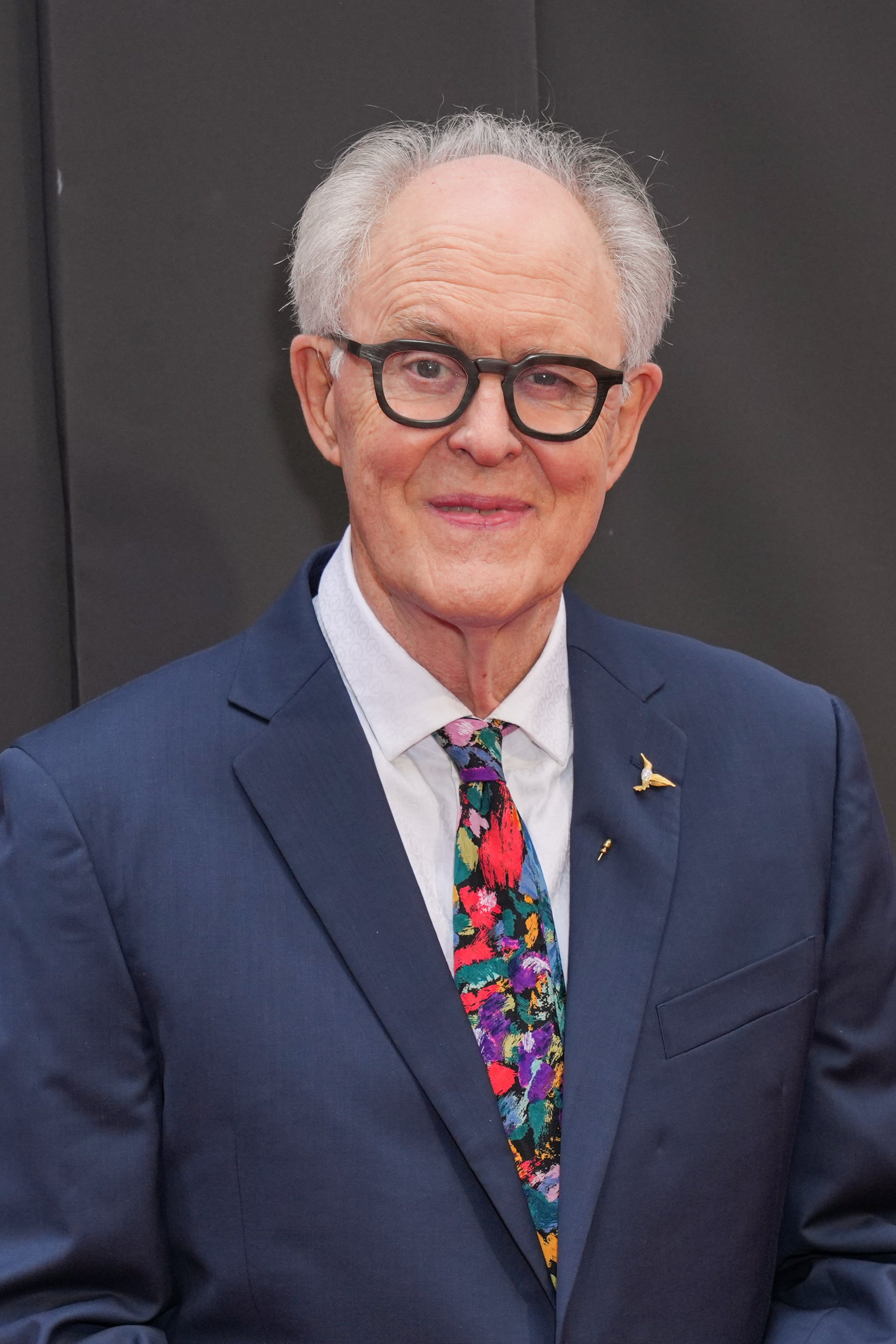John Lithgow in a suit with and printed tie and glasses stands on a red carpet, smiling warmly for the camera
