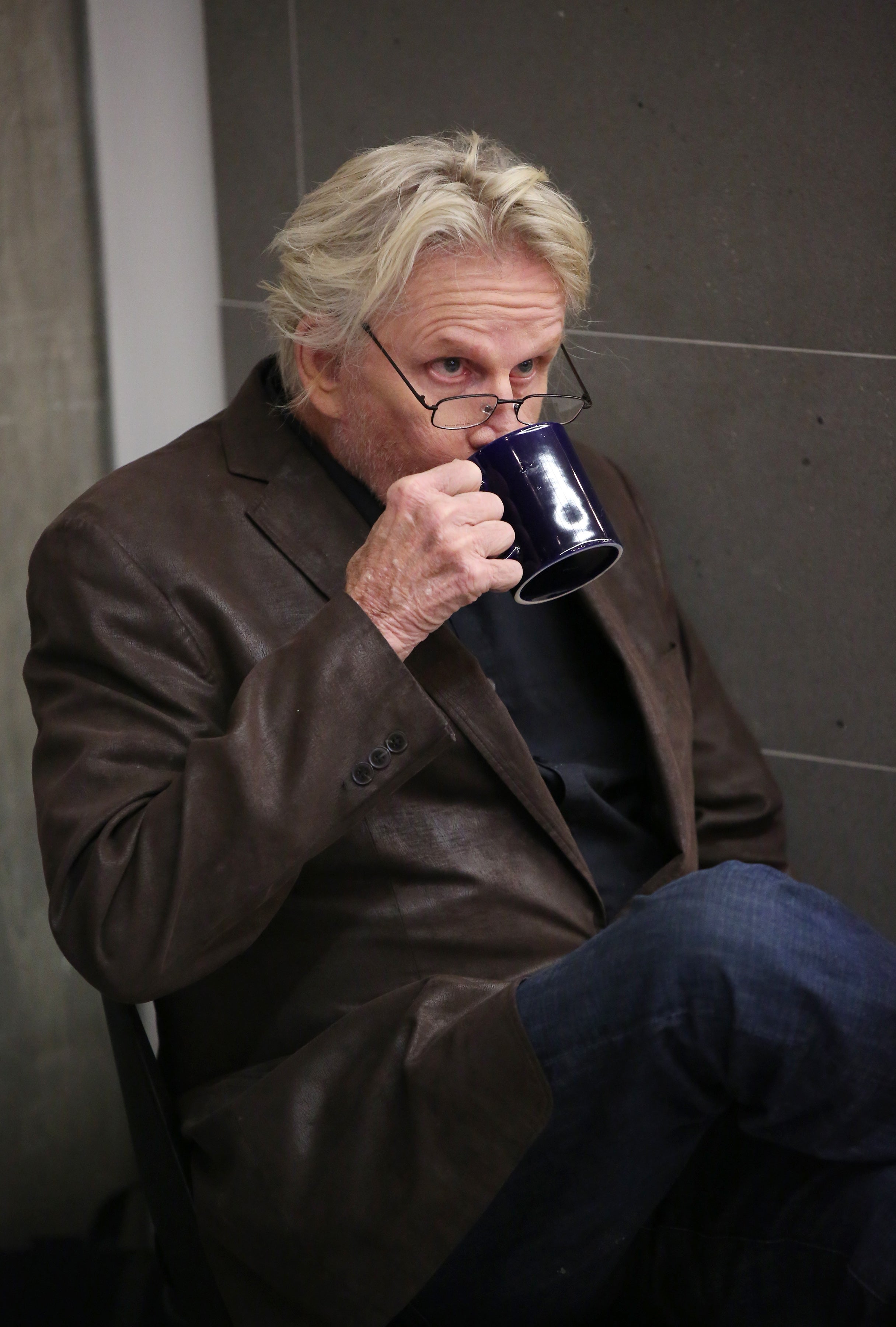 Gary Busey with glasses and casual attire, sipping from a mug while seated