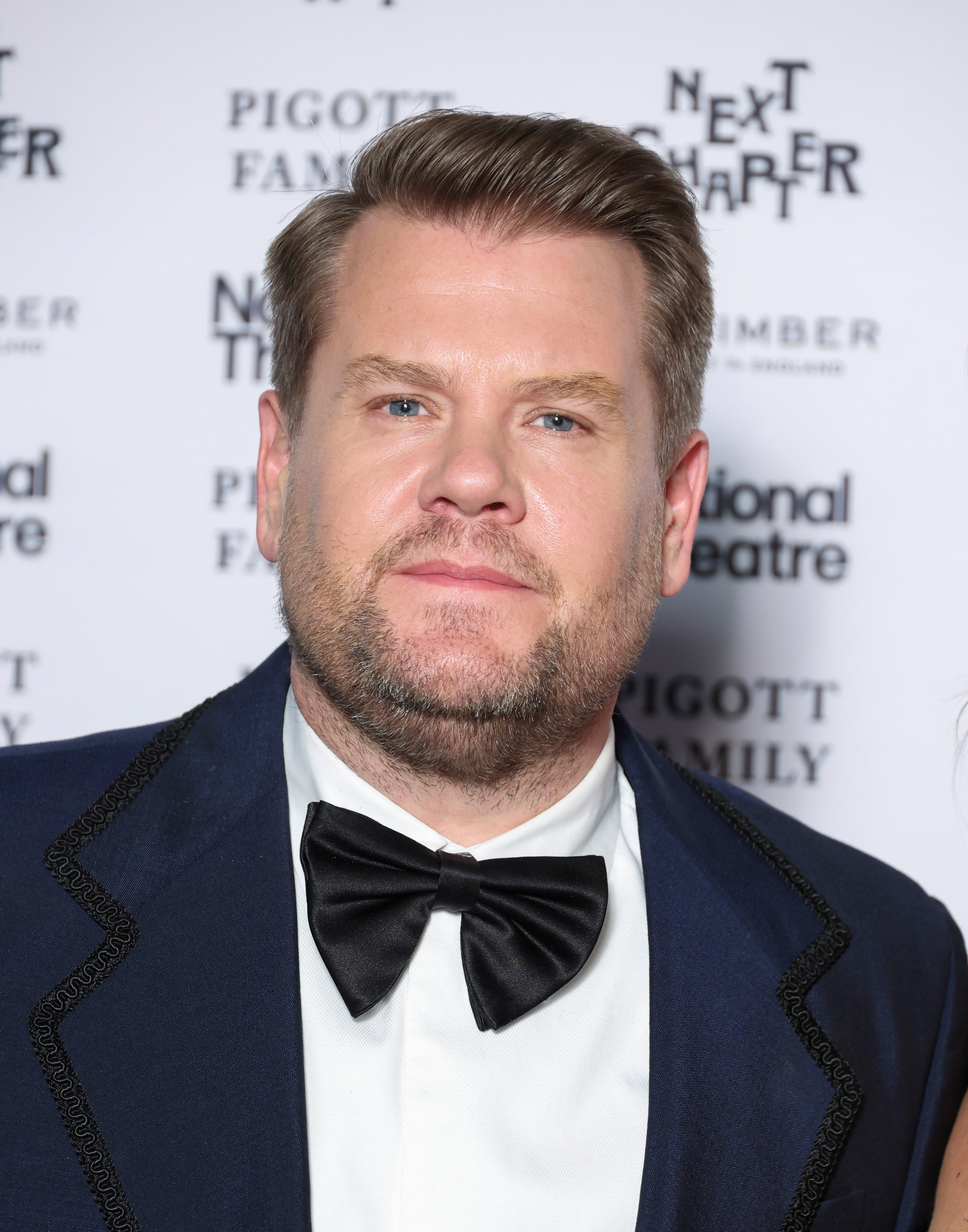 James Corden in a suit with a bow tie at a theater event backdrop with sponsor logos