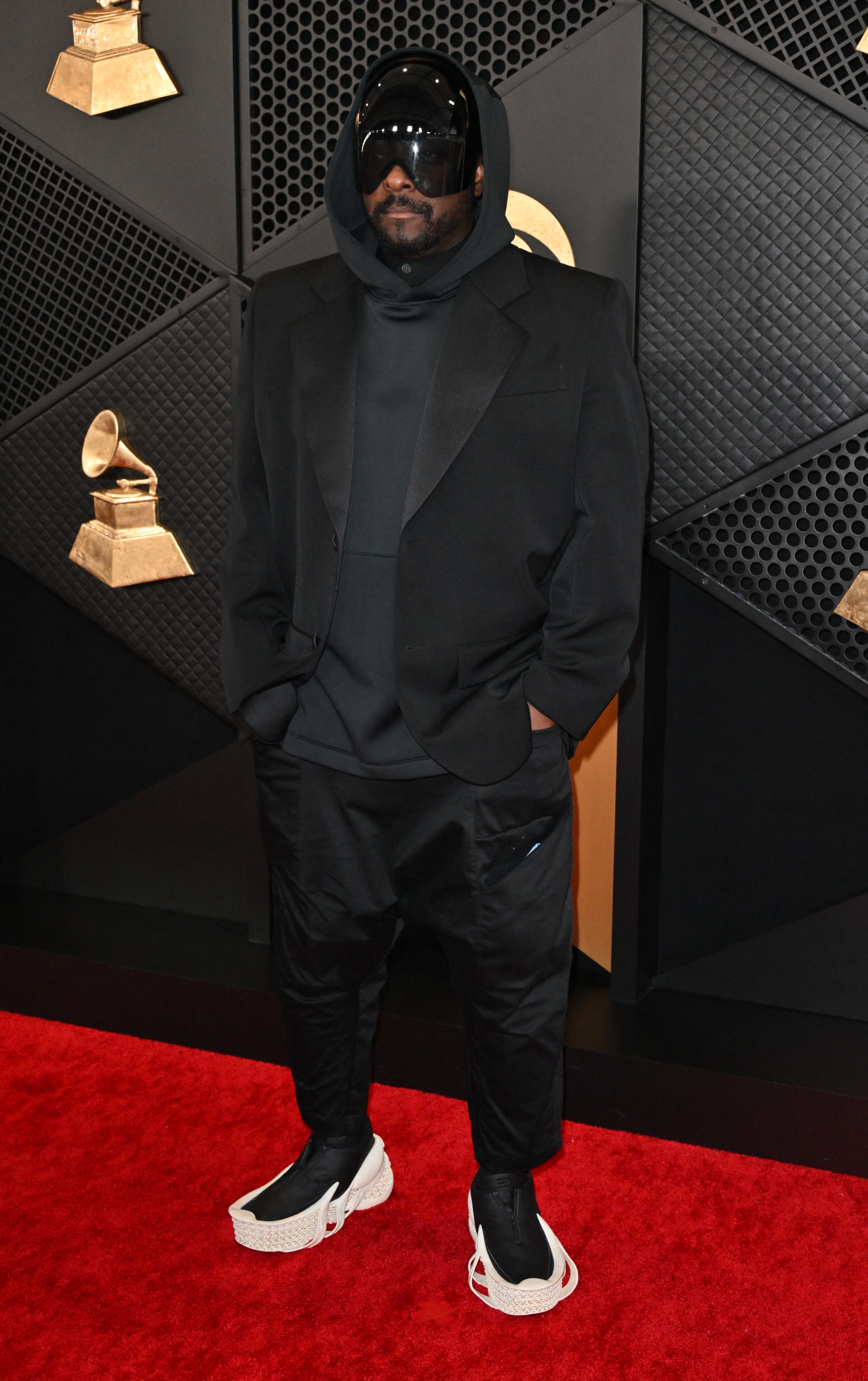 will i am on red carpet in an oversized hoodie, blazer, pants, and large visor-style sunglasses, with futuristic sneakers