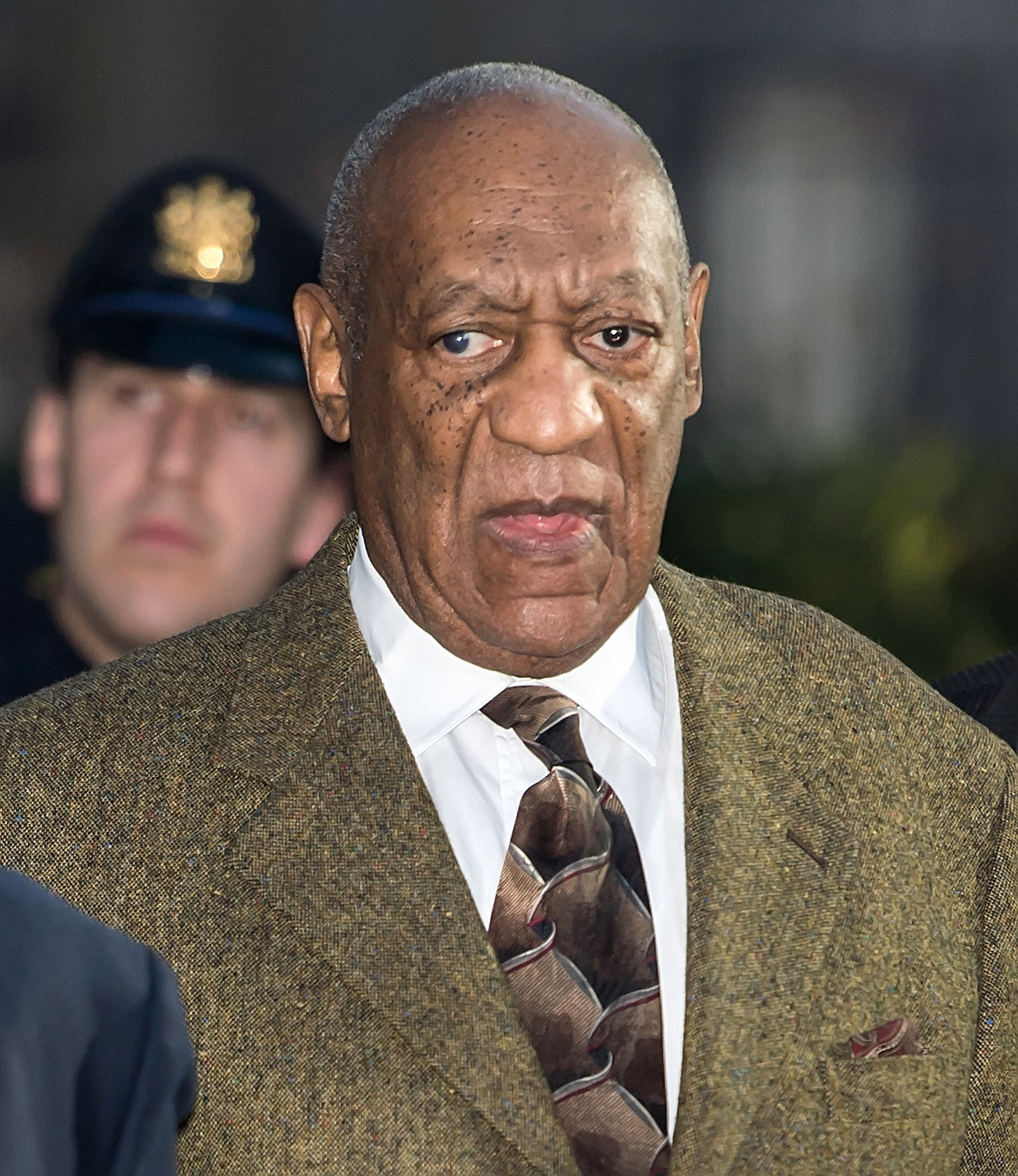 Bill Cosby in a patterned tie and suit outside, with a blurred security figure in the background