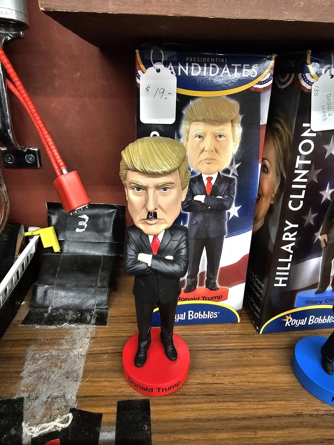 A bobblehead of a figure in a suit with crossed arms and a distinctive hairstyle is on display. The box has text referencing "Donald Trump."
