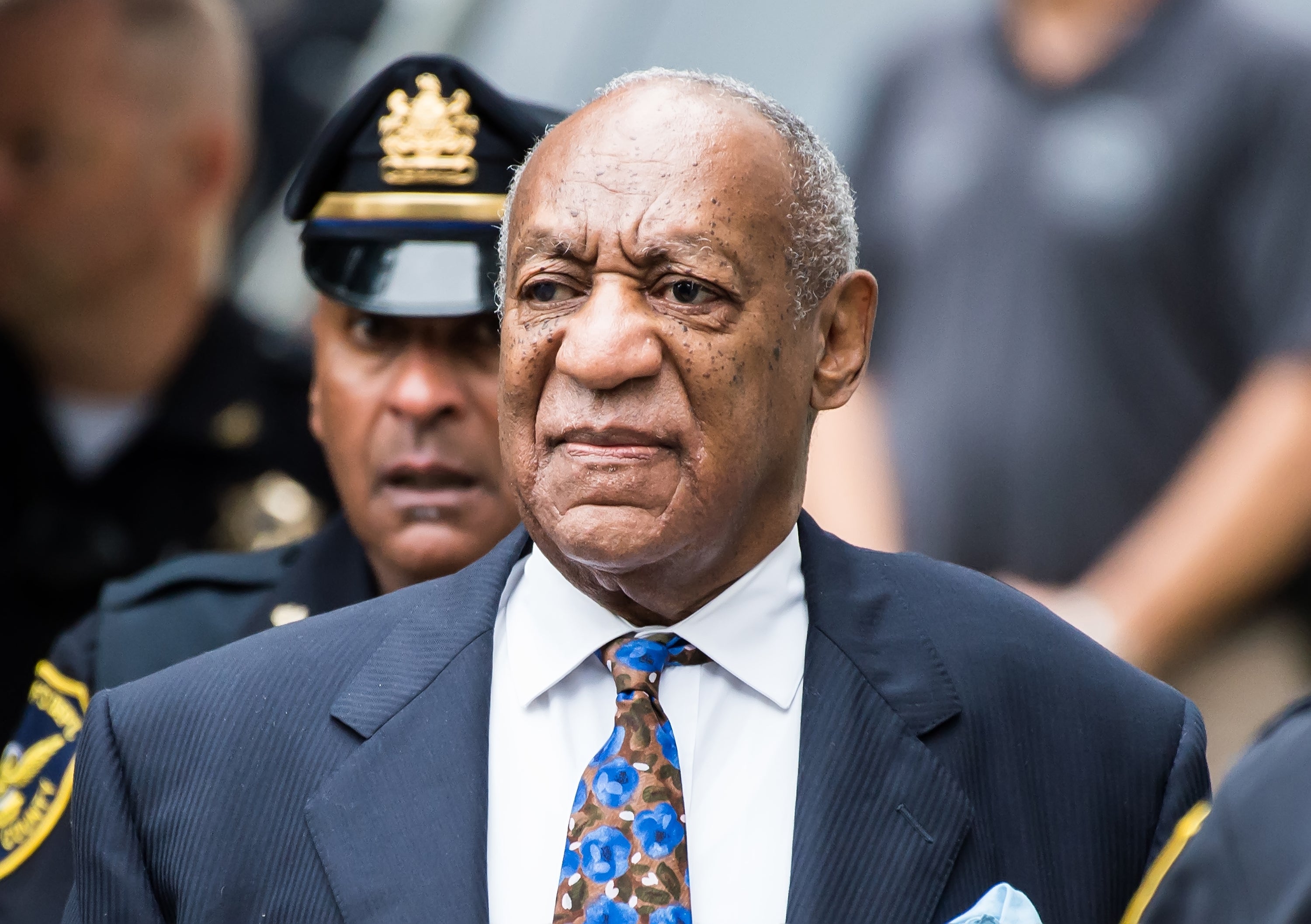 Bill Cosby in a suit with a floral tie, surrounded by security personnel