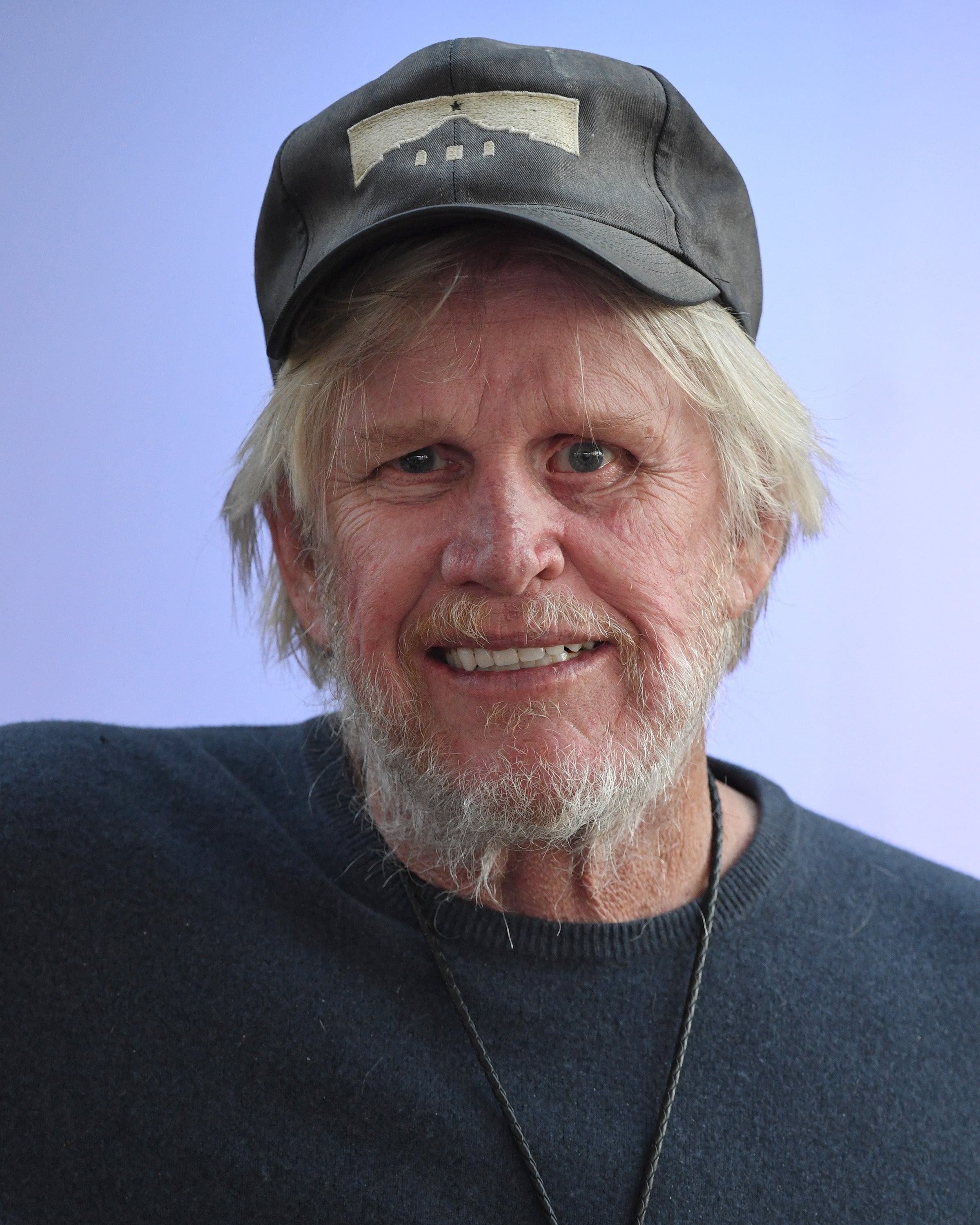 Gary Busey smiling, wearing a casual cap and sweater, with a light beard and mustache, set against a simple background