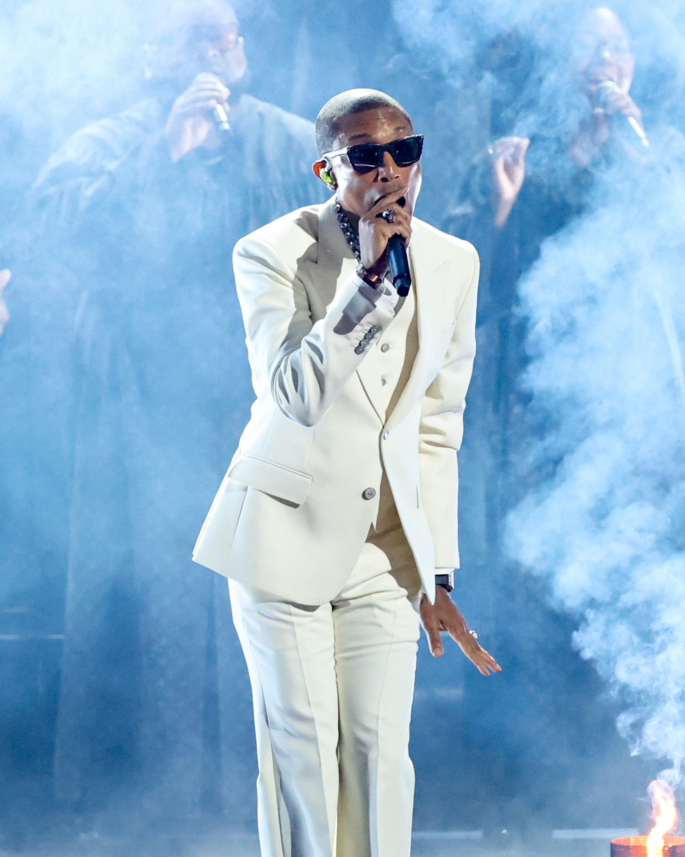 Pharrell in a stylish suit and sunglasses performs on stage, surrounded by smoke effects