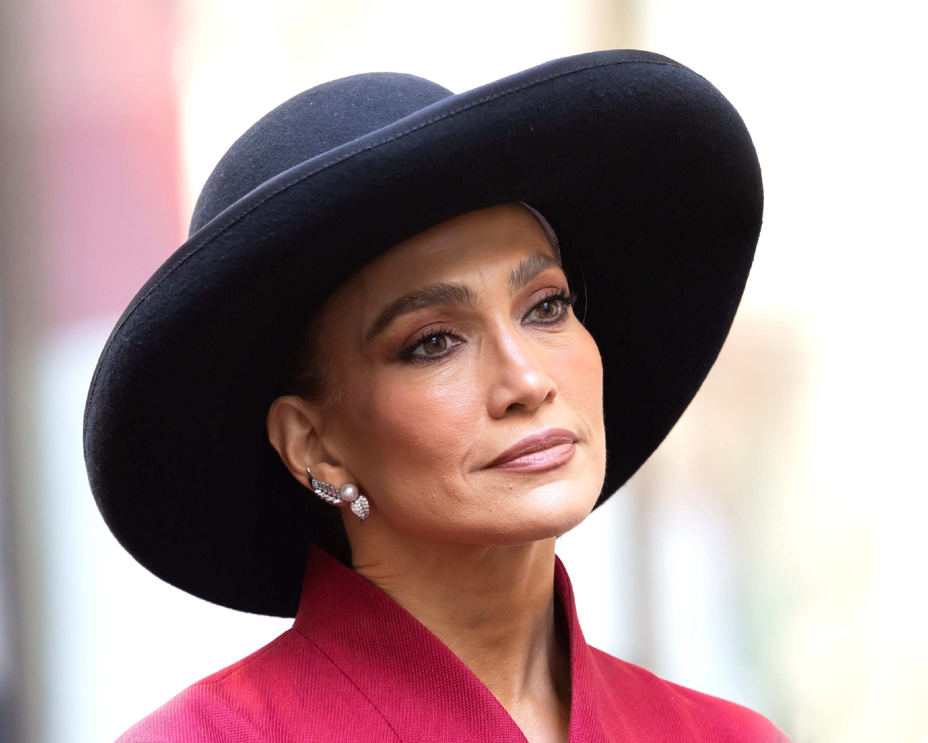 Jennifer Lopez wearing a wide-brimmed hat and a stylish suit with statement earrings, looking thoughtful