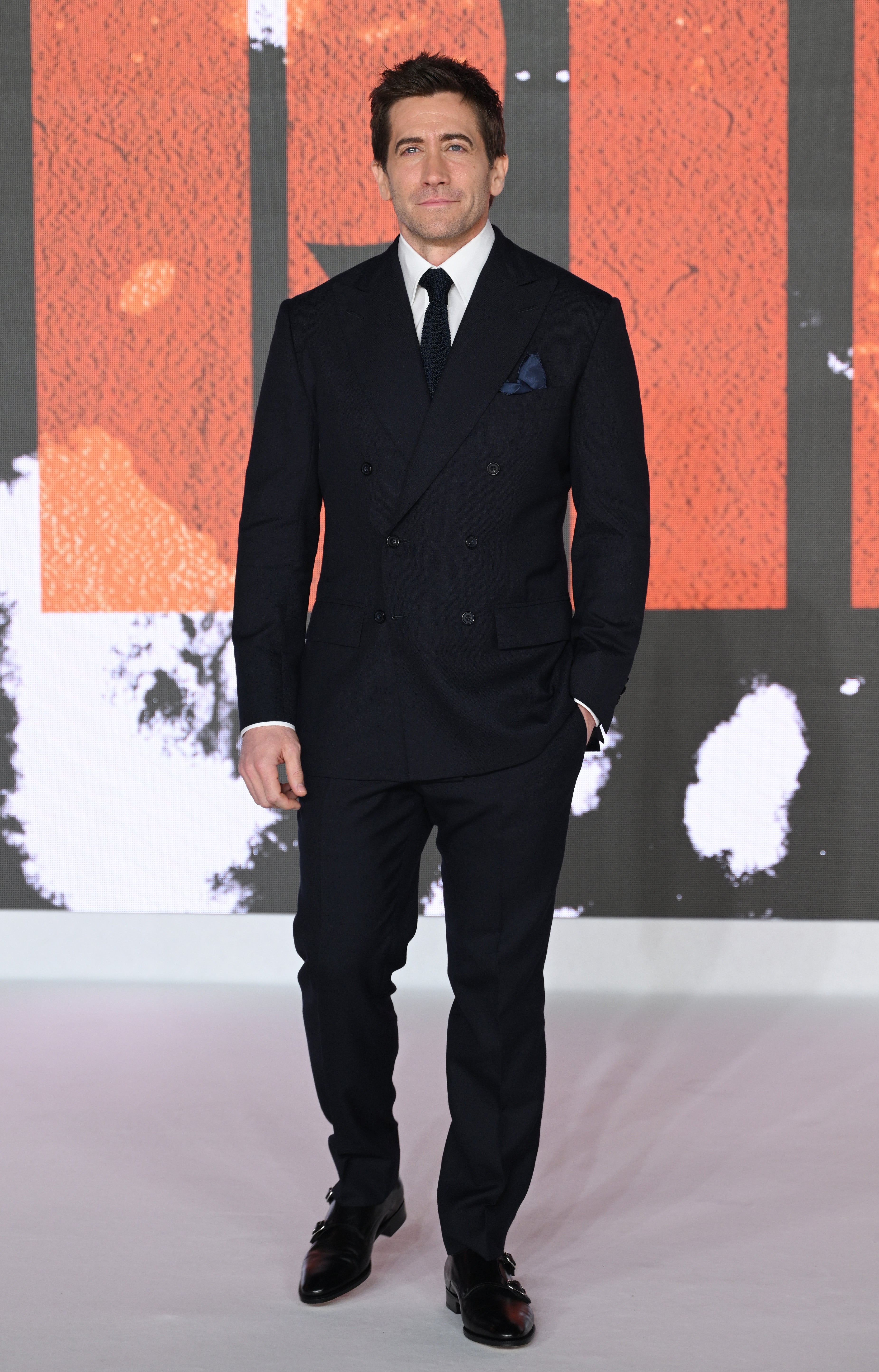 Jake Gyllenhaal in a tailored suit on a red carpet, standing confidently with one hand in pocket, against a textured backdrop