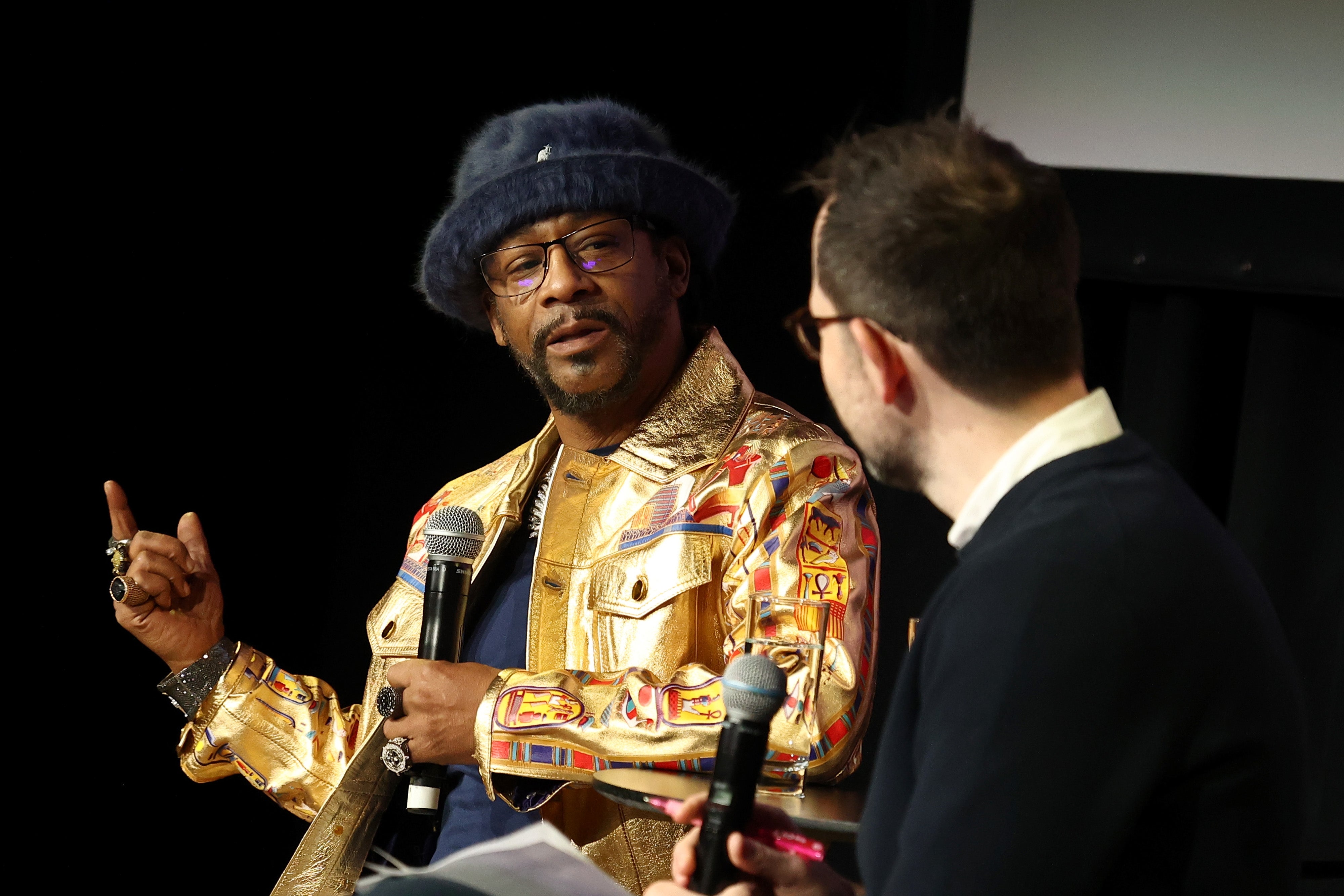 Katt Williams in a patterned jacket and fluffy hat speaks into a microphone on stage, gesturing with a hand, while another person listens attentively